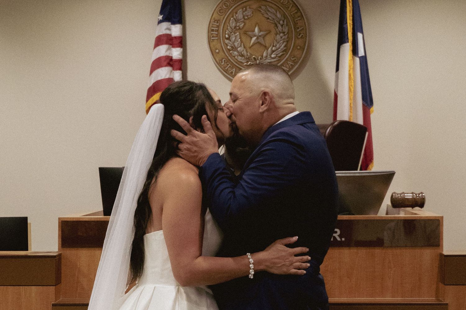 A couple shares their first kiss during a courthouse wedding ceremony with an American flag and Texas seal visible.