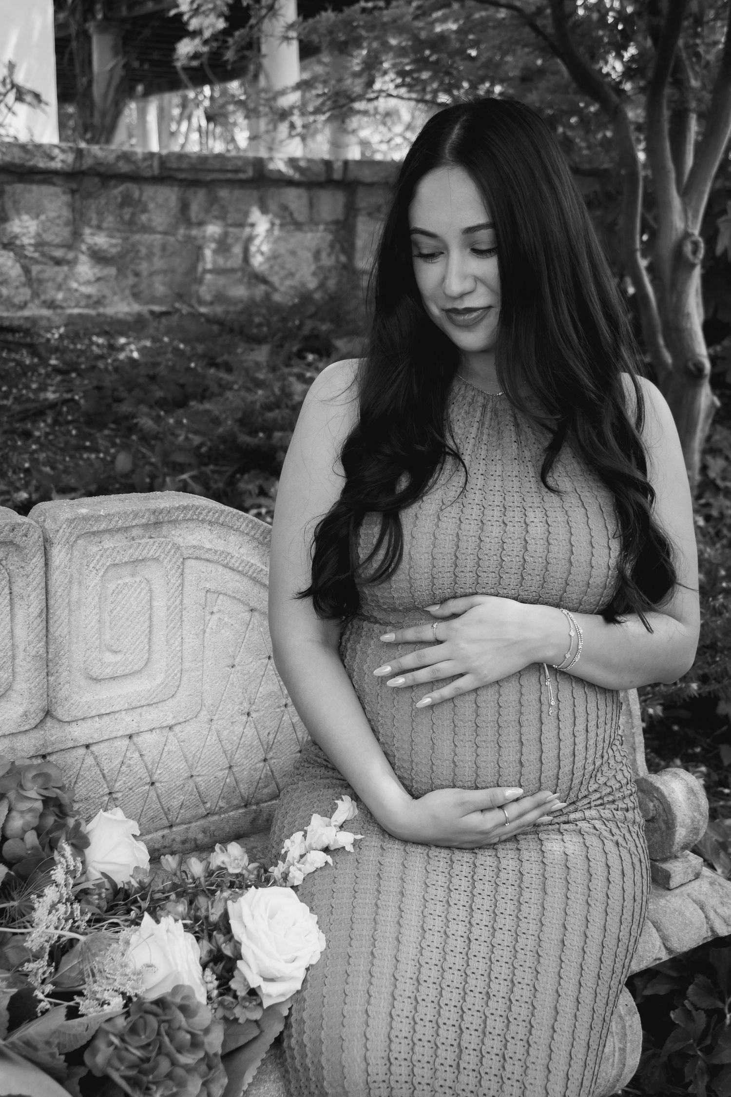 Black and white maternity photo of woman in knit dress standing by stone wall with roses in a garden setting.