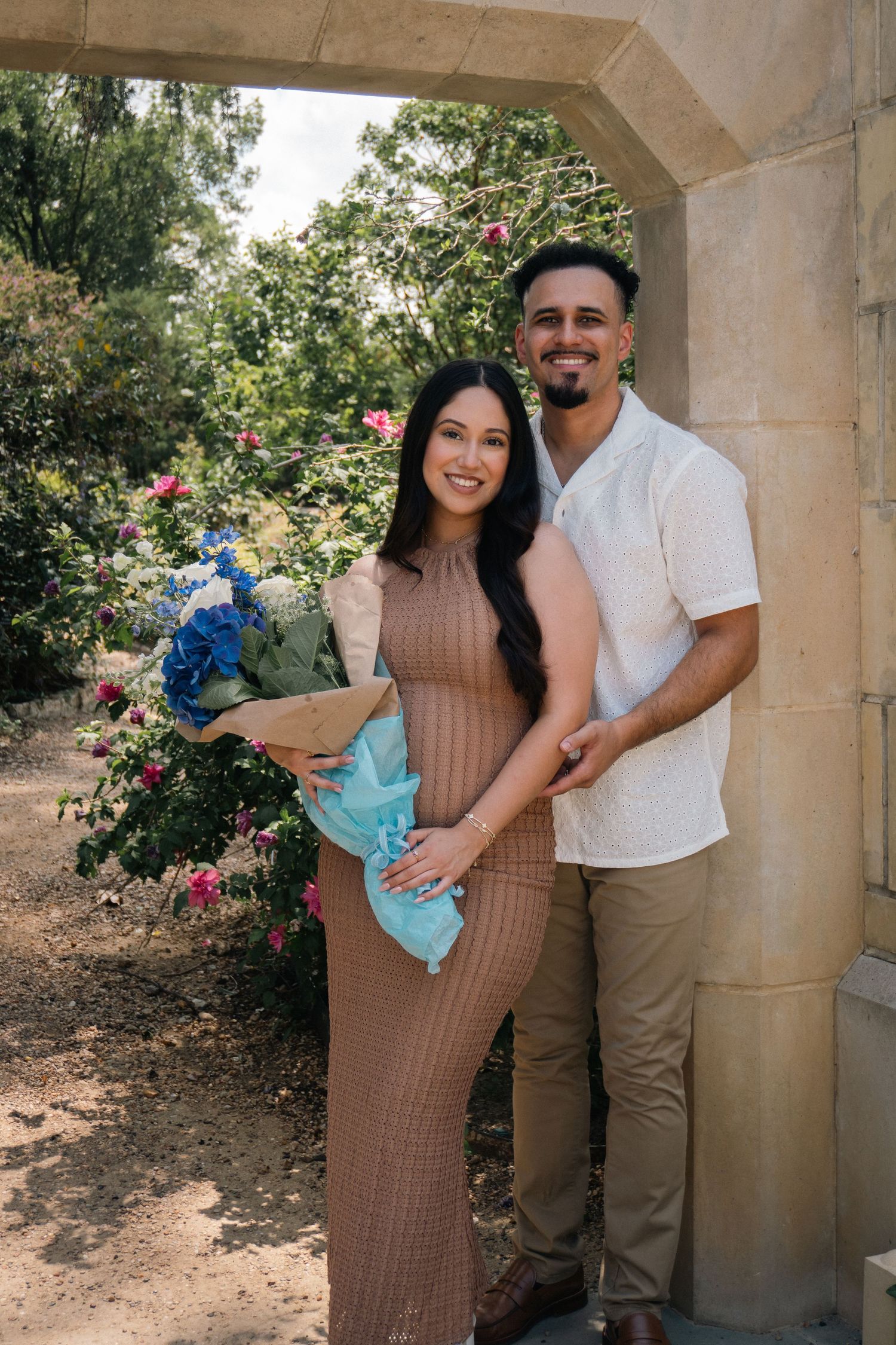 A couple poses together under a stone archway in a garden setting with blooming flowers.