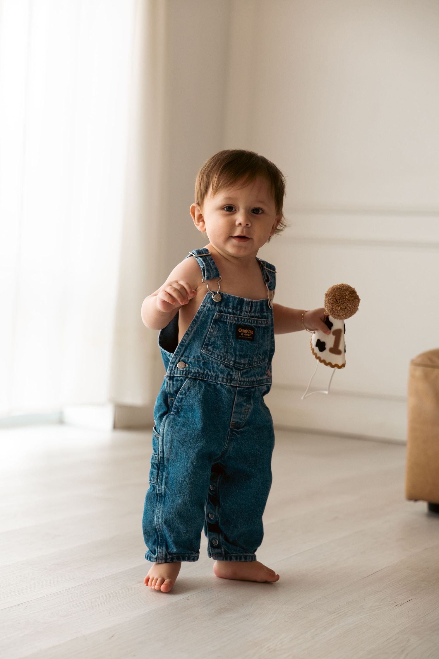 Toddler wearing blue denim overalls practices walking on hardwood floor in sunlit white room.