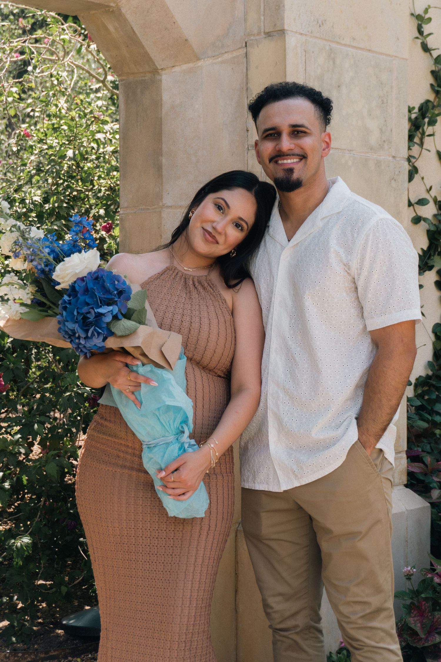 A couple poses together outdoors against a stone archway, with the woman holding a bouquet of blue flowers.
