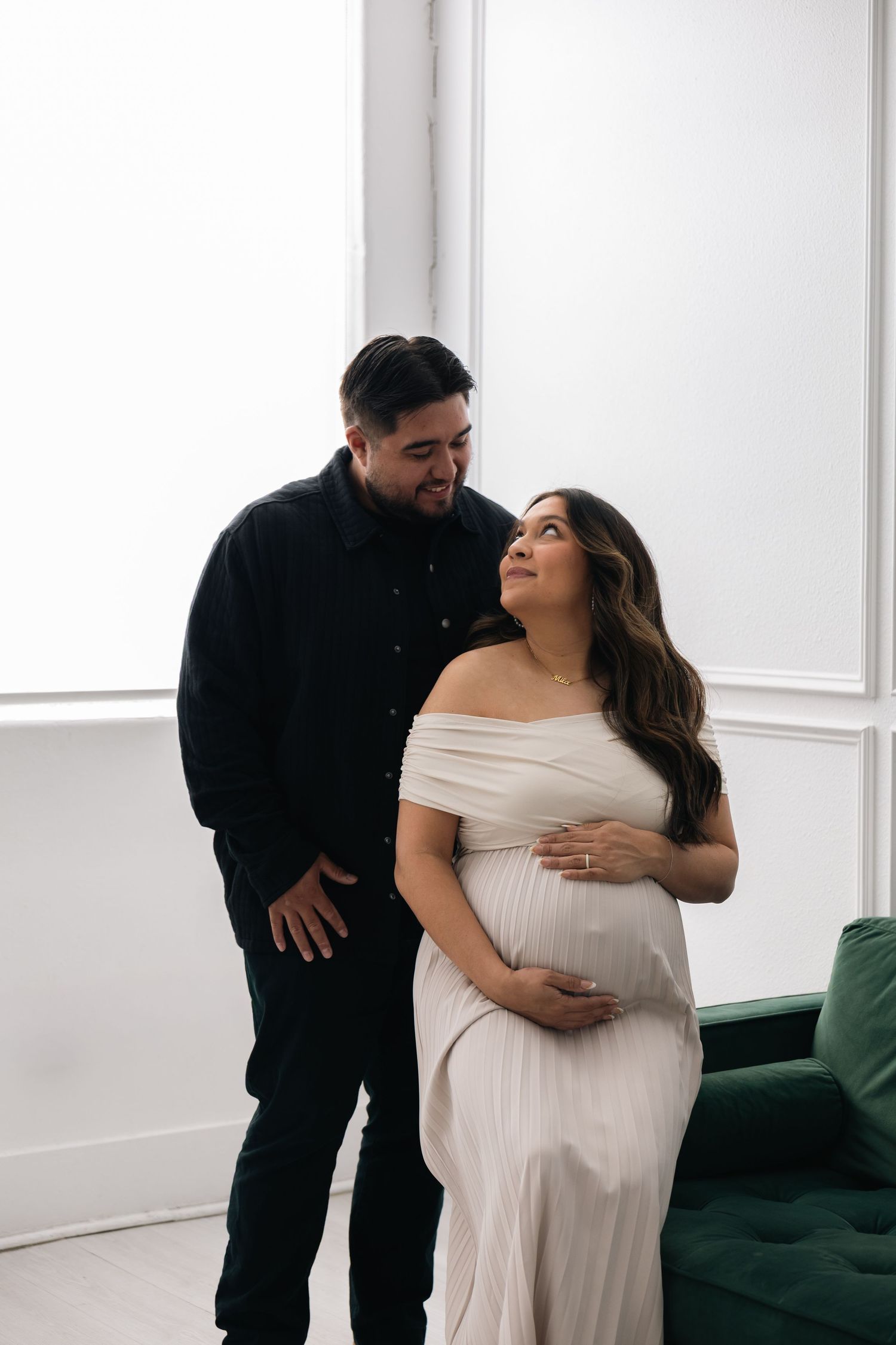 A couple shares an intimate moment in a minimalist white room with a pregnant woman wearing an off-shoulder cream dress.