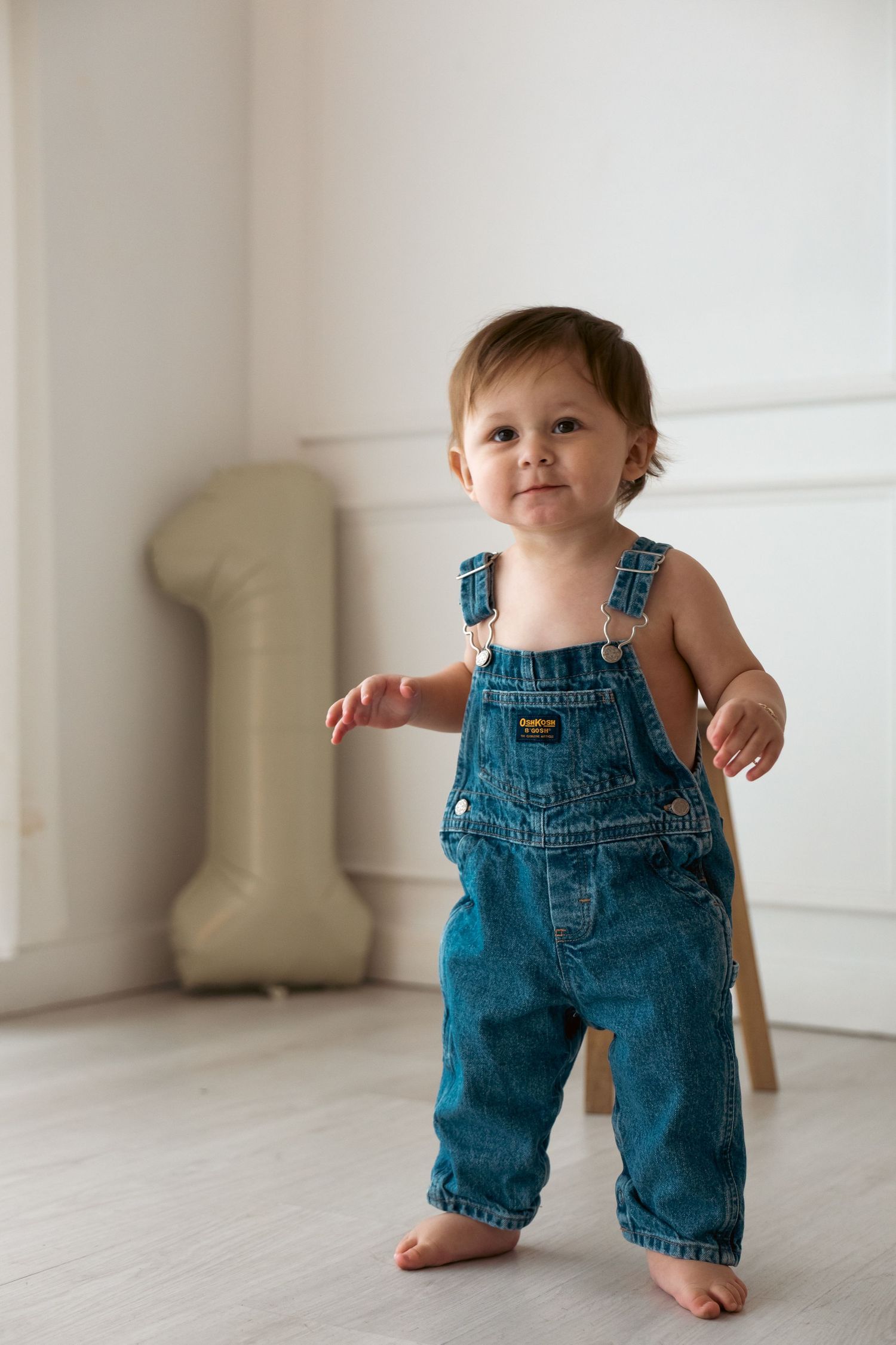 Toddler in denim overalls taking first steps on white wooden floor.