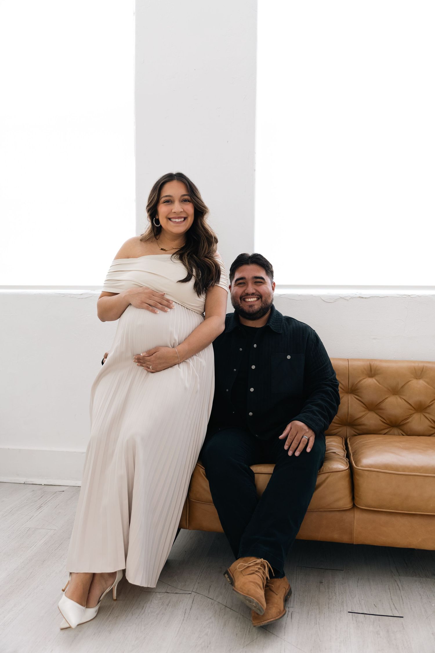 A couple poses together on a tan leather couch, with the woman wearing an off-shoulder cream dress and the man in dark attire.