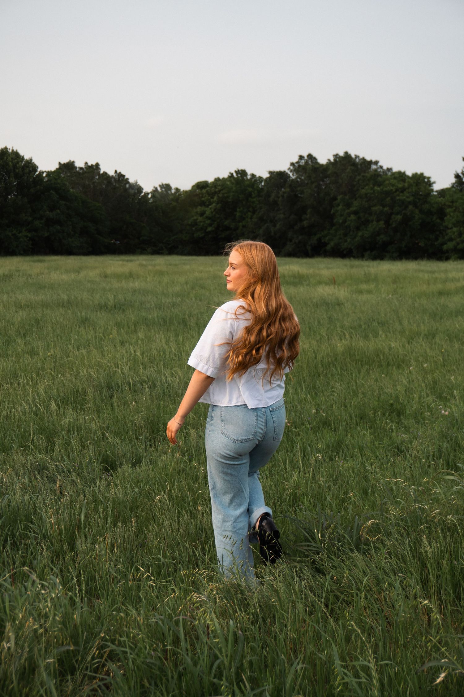Person with long wavy hair wearing jeans and white shirt walks through tall grass in a serene meadow at dusk.