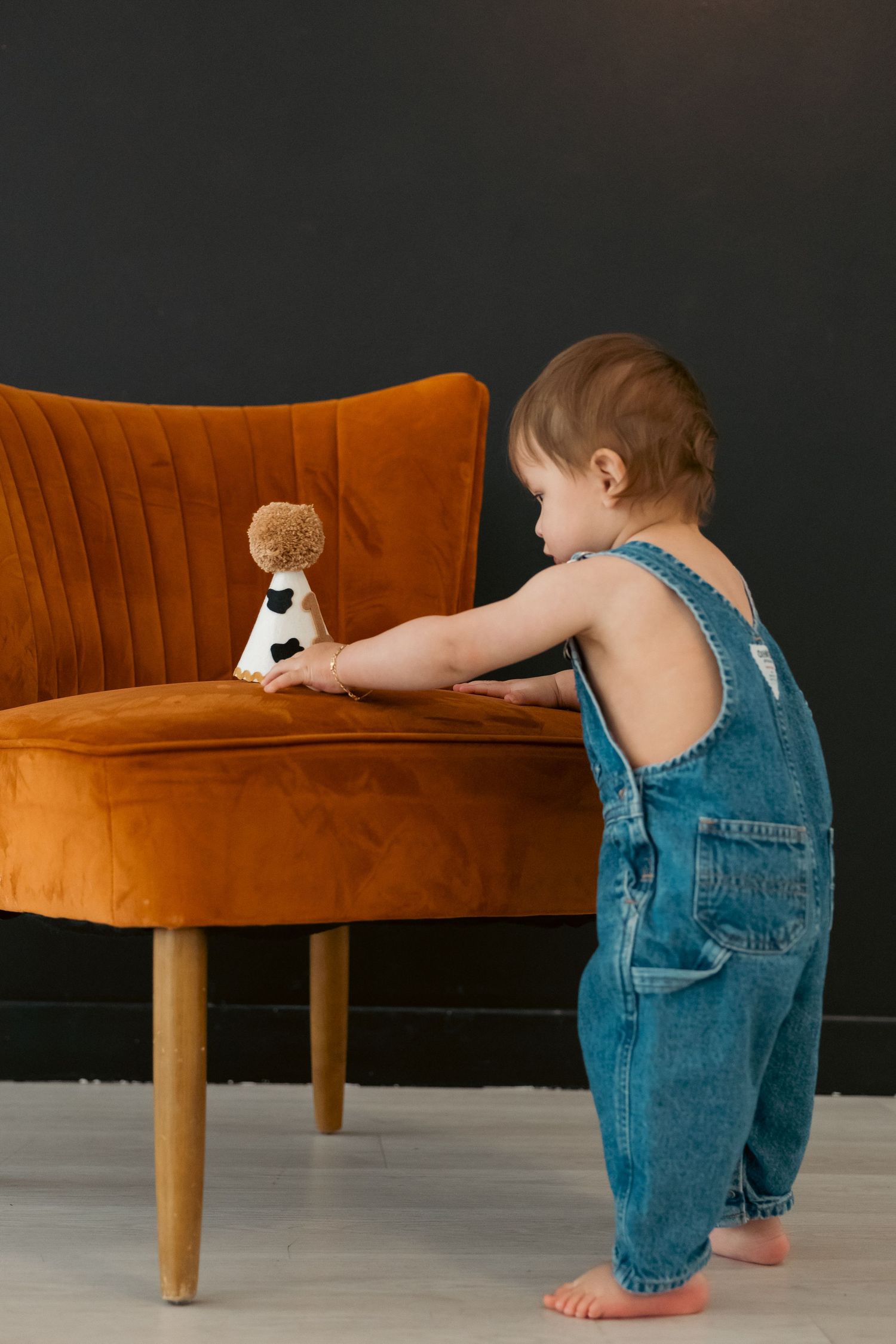 A toddler in denim overalls reaching towards an orange velvet couch in a minimalist setting.