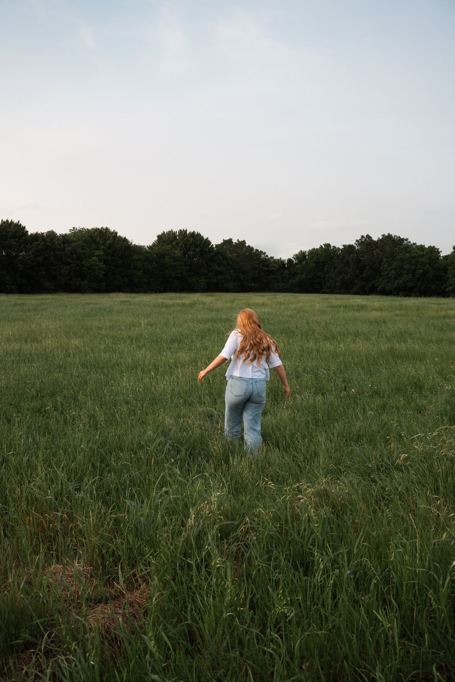 A person with long blonde hair wearing jeans walks through a lush green meadow at dusk with trees in the distance.