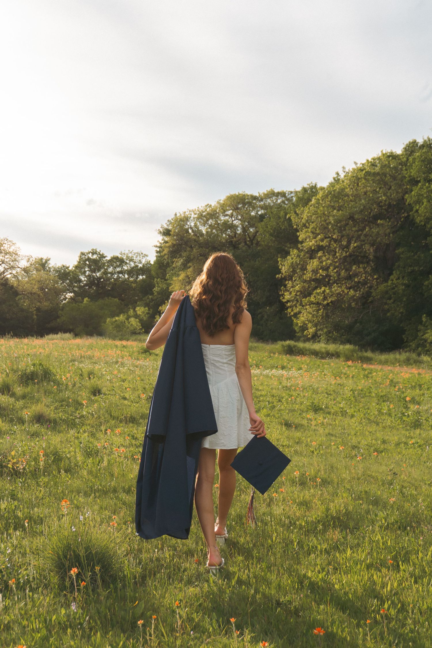 A person in a white dress carries a graduation gown across a sunny hillside meadow at golden hour.