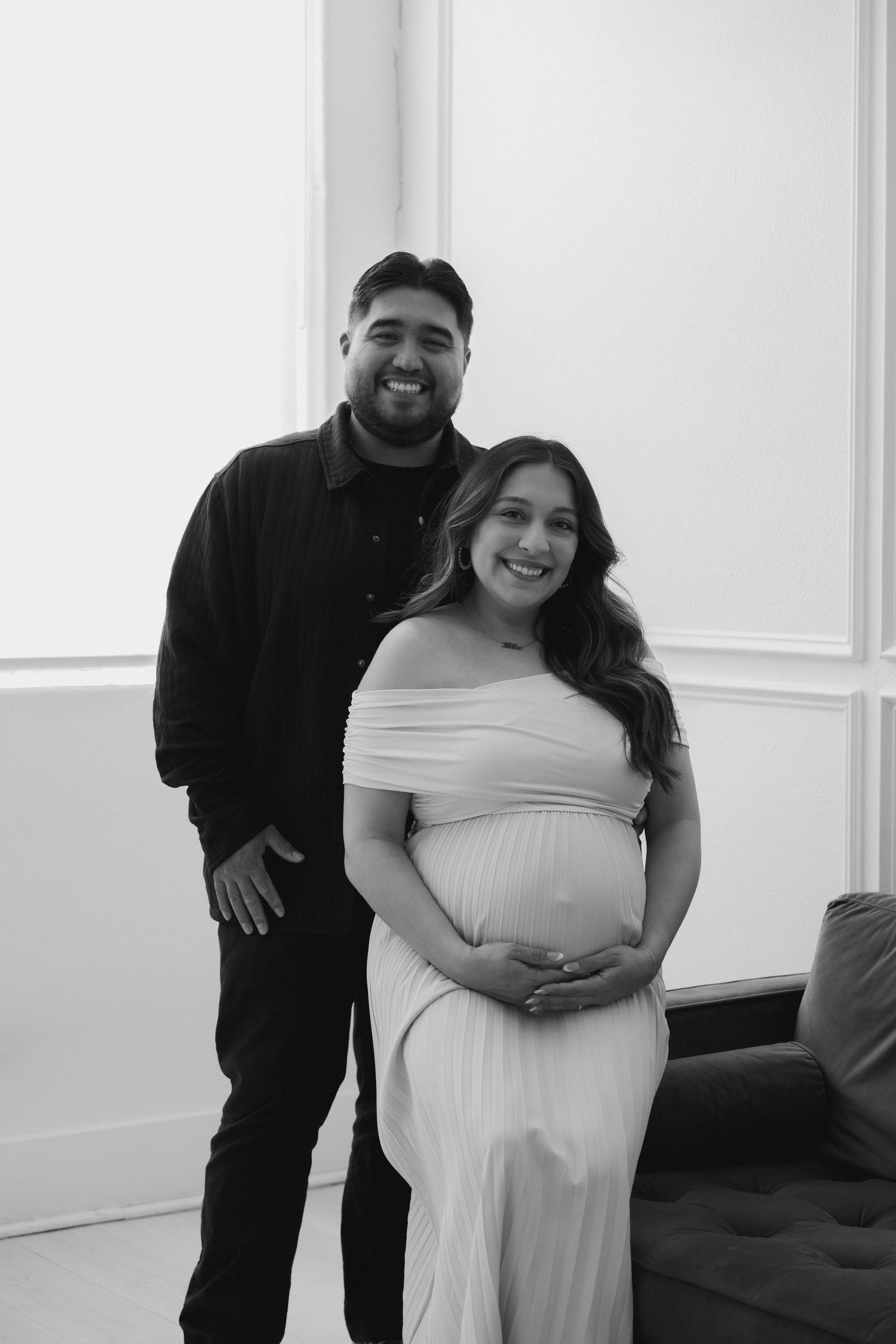 A couple in black and white poses together, with the woman wearing a white off-shoulder maternity dress.