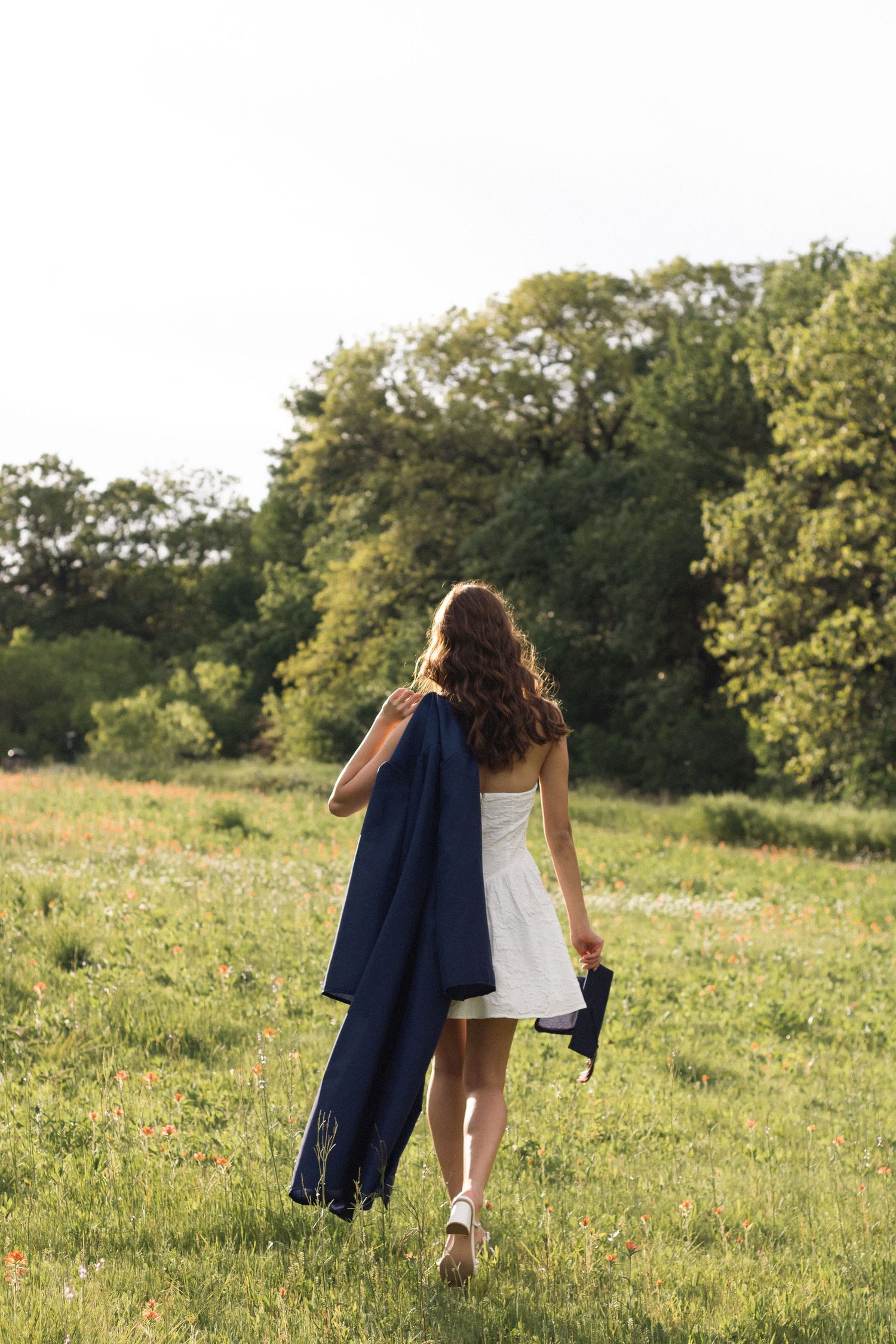 Person in white dress walks through sunny meadow carrying navy blue jacket on summer afternoon.