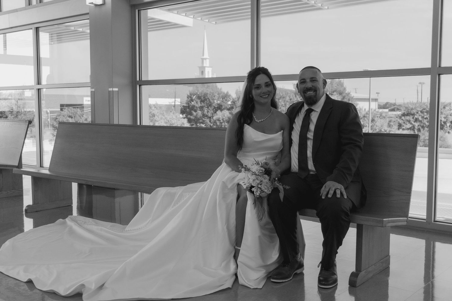 A couple in wedding attire sits together on a bench by large windows in black and white.