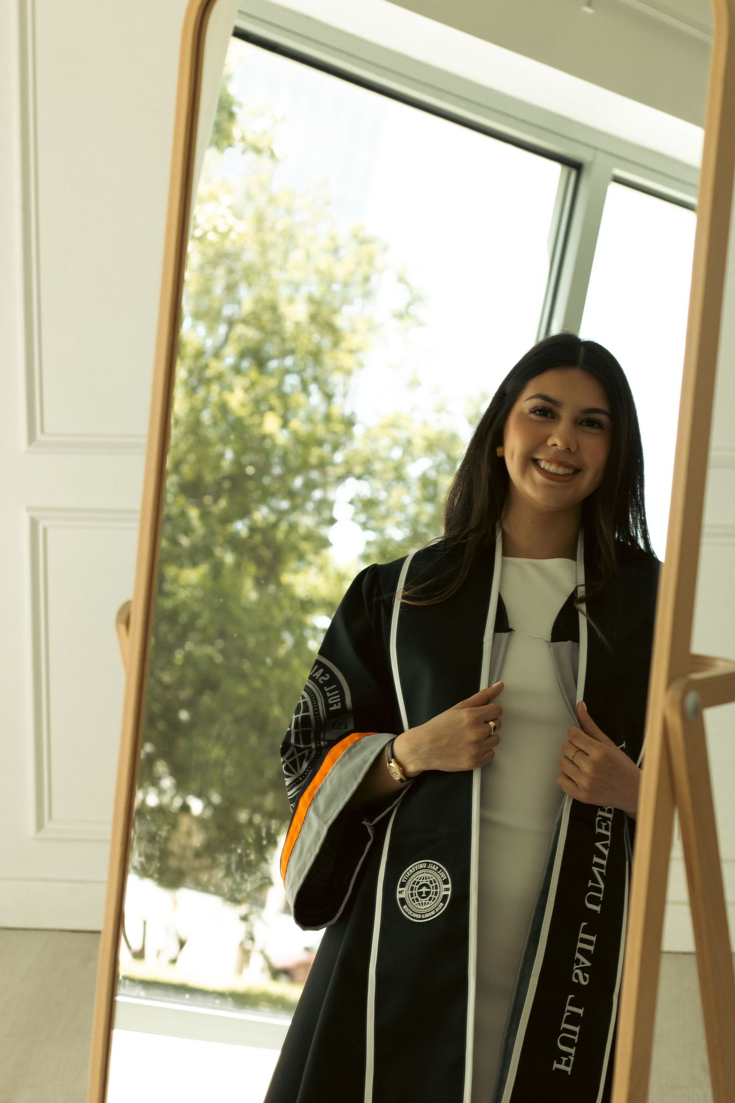 Graduate holding diploma and posing in academic regalia by window.