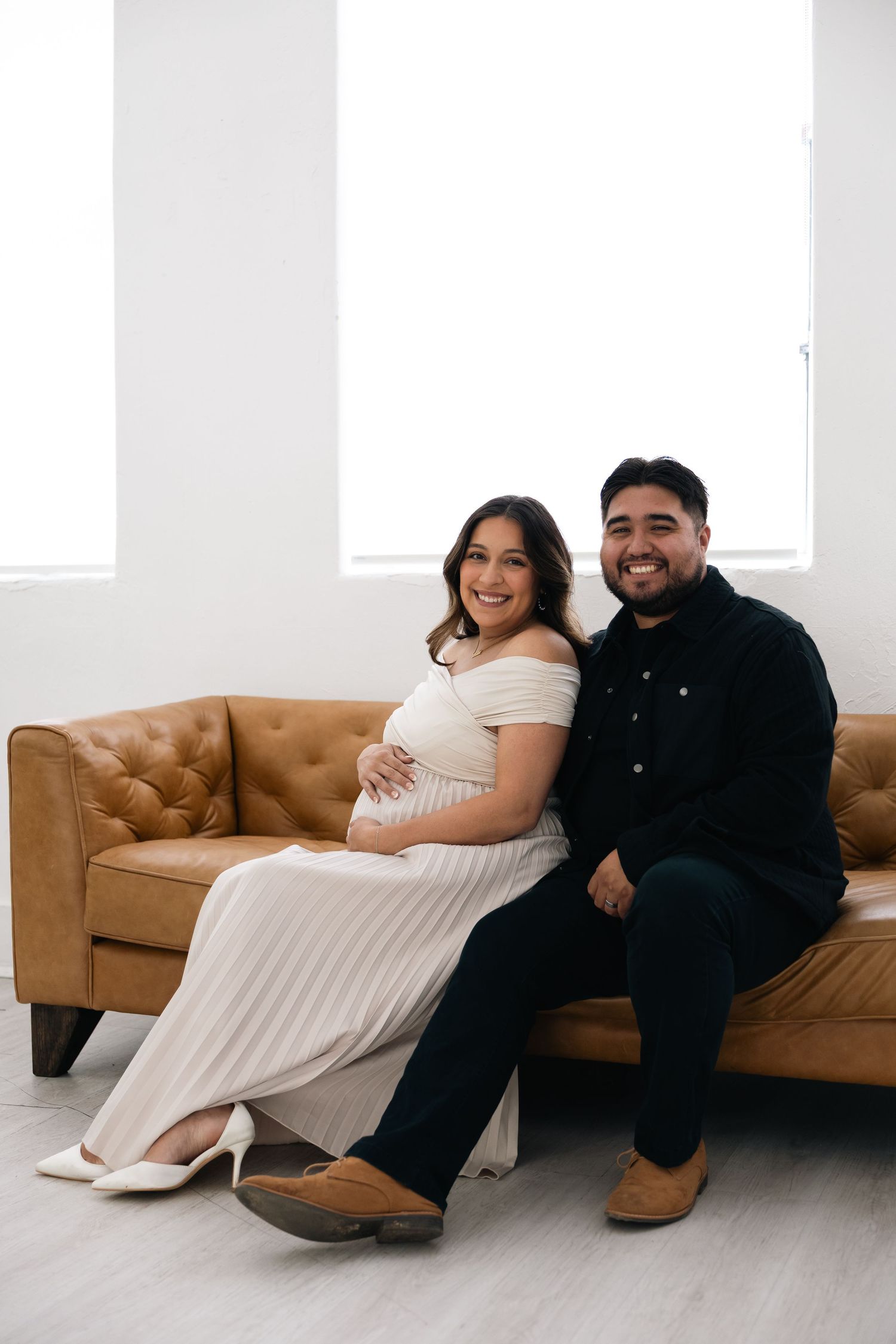 A couple sits together on a brown leather couch in a bright white room during a photo session.