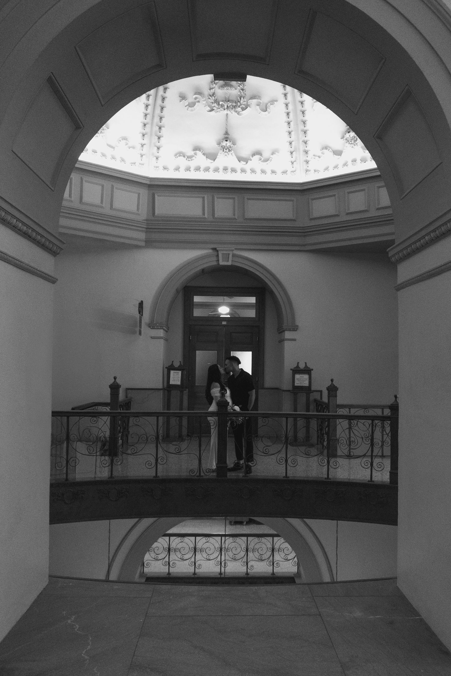 A silhouetted couple stands on a balcony inside an ornate domed building with classical architecture.