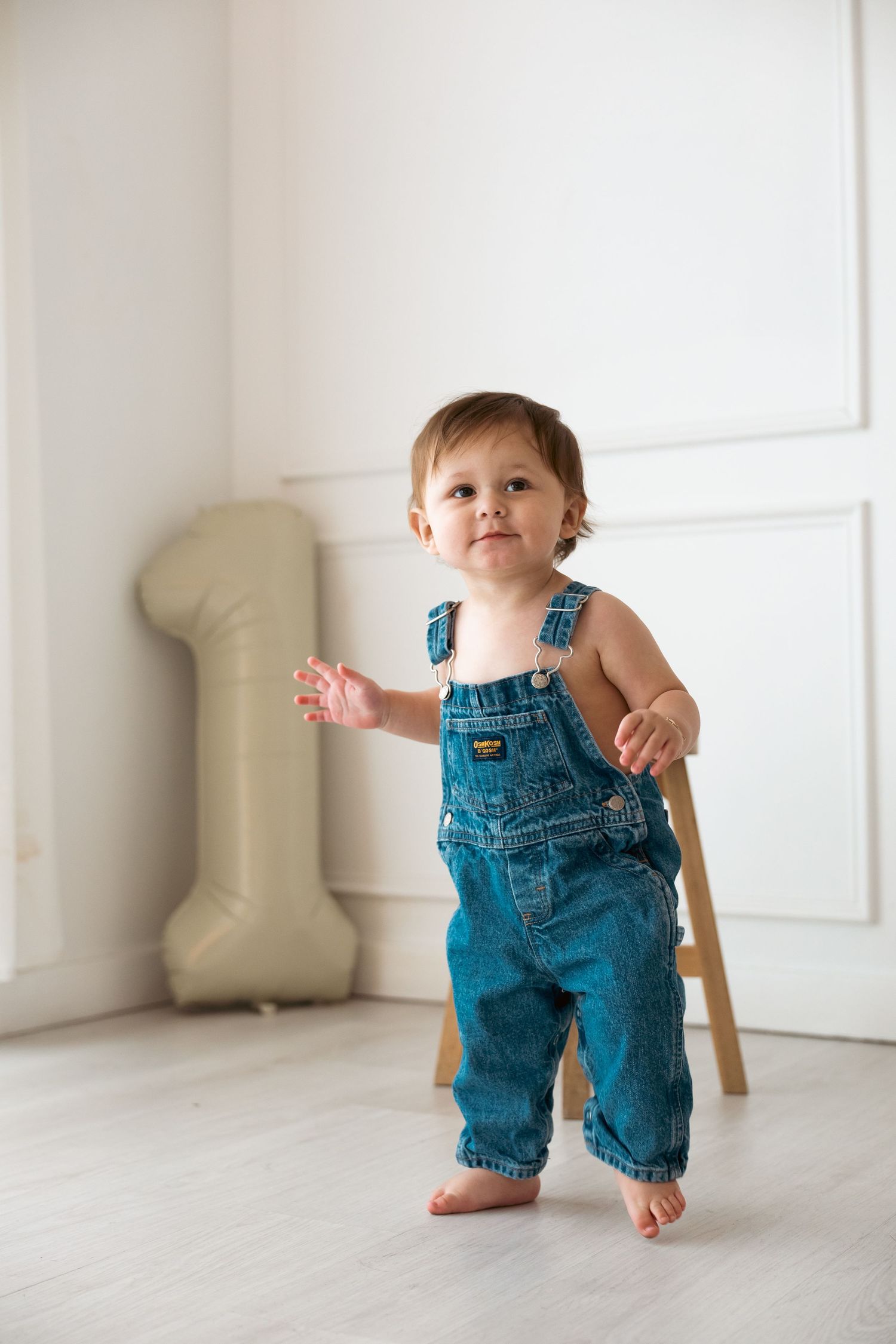 Small toddler in denim overalls taking first steps on light wooden floor in bright minimalist room.