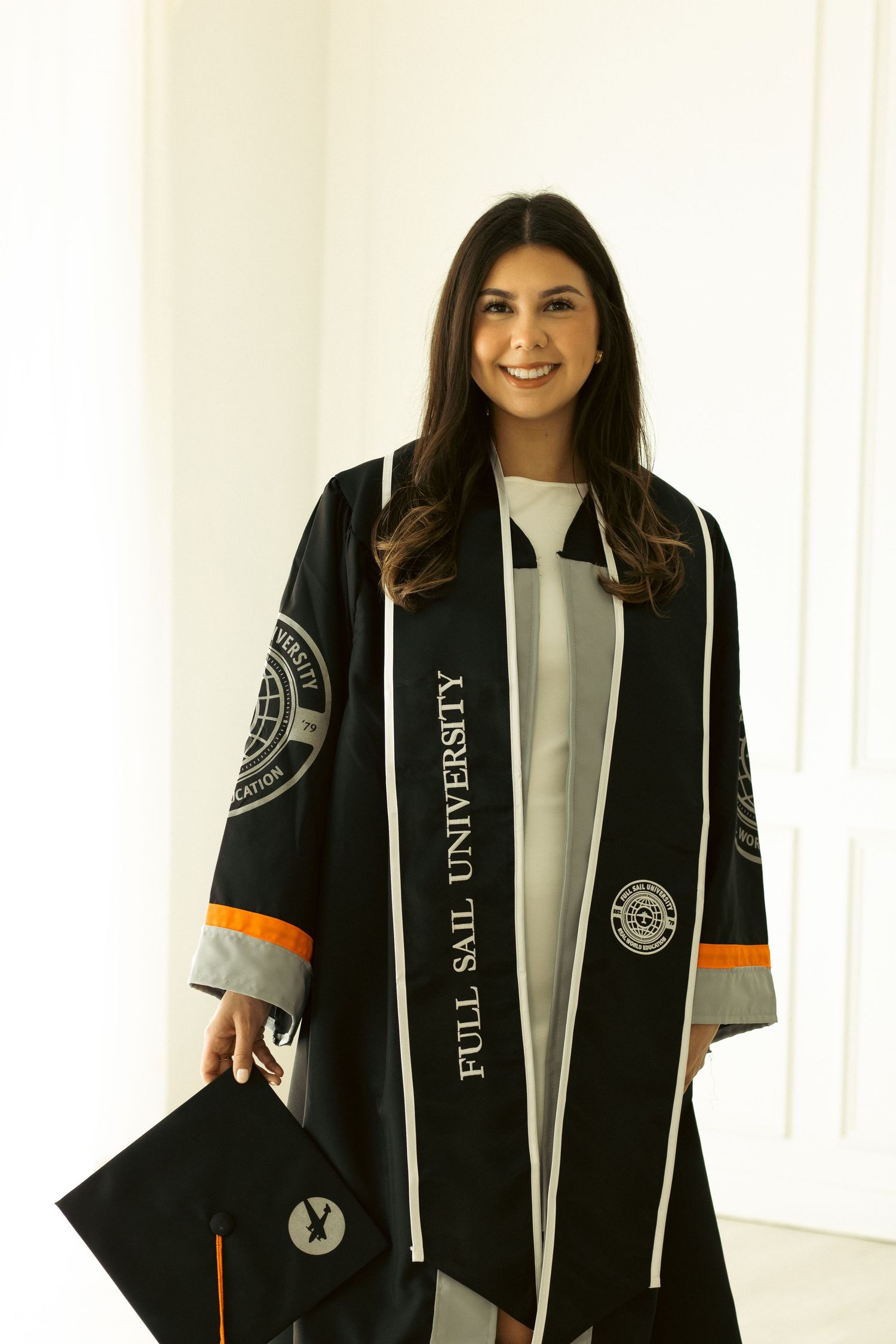 Graduate posing in black graduation gown with orange accents against white backdrop.