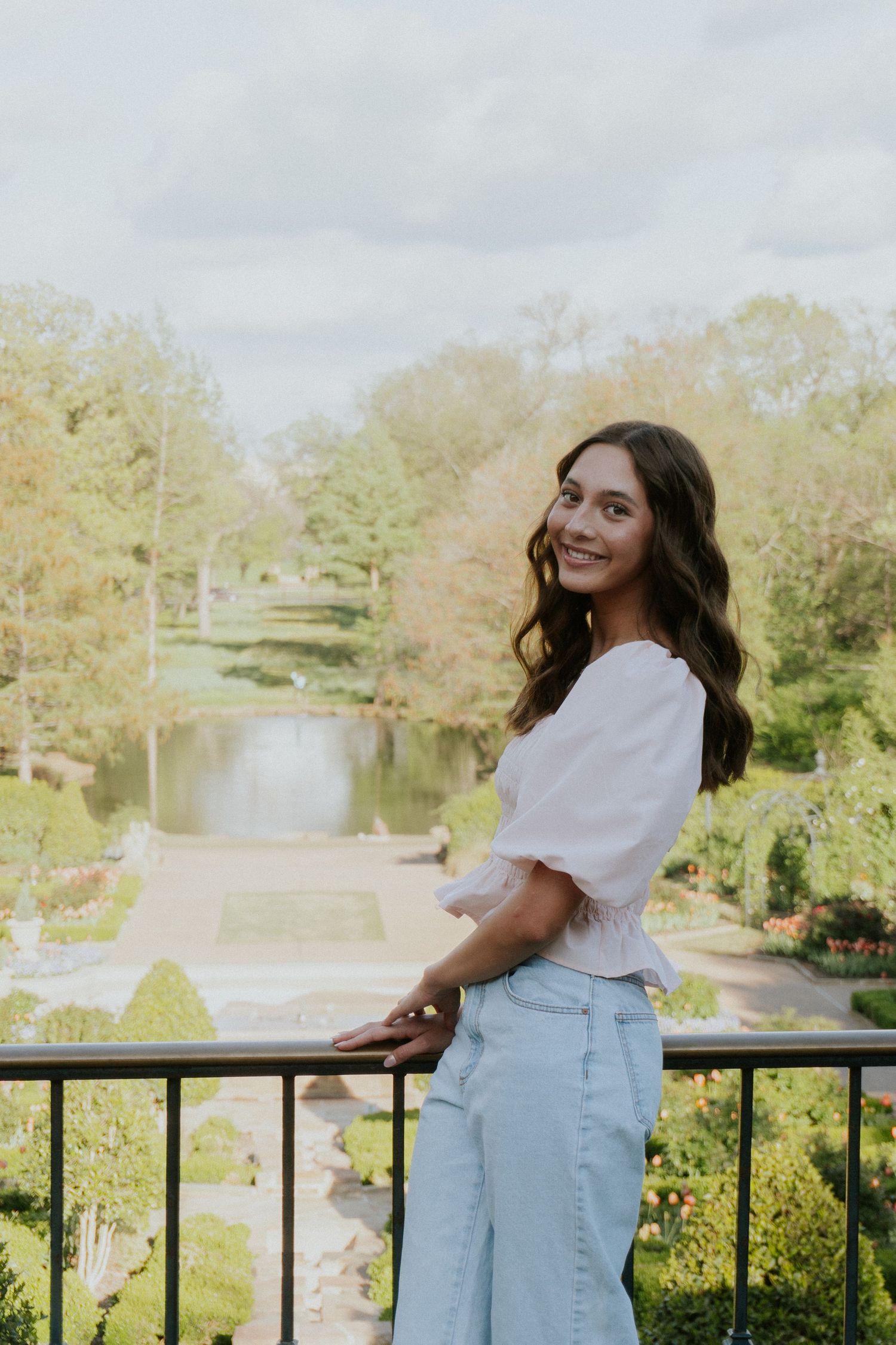 Person in white top and jeans posing by a railing overlooking a scenic pond.