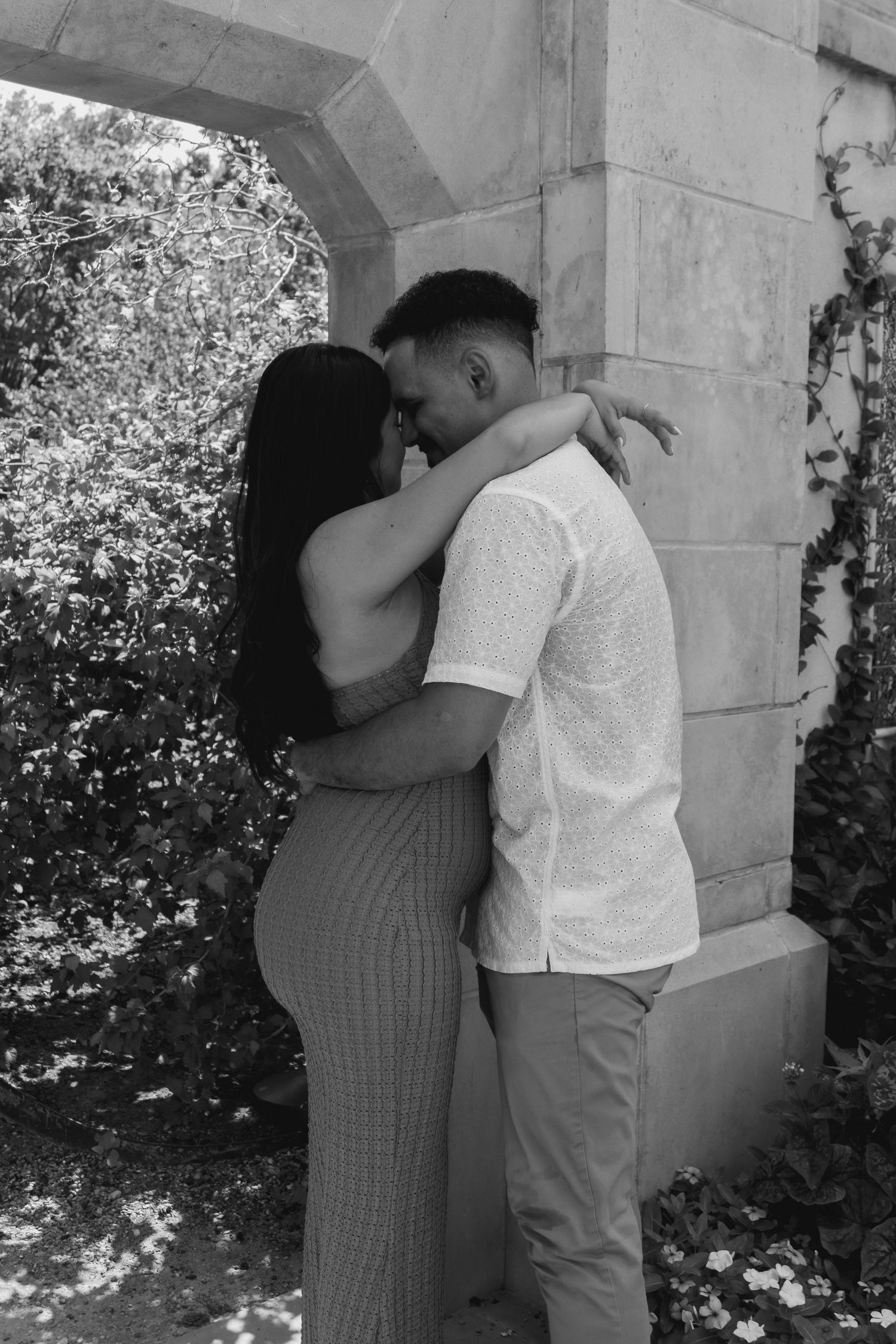 A romantic couple embraces under a stone archway in a garden setting.