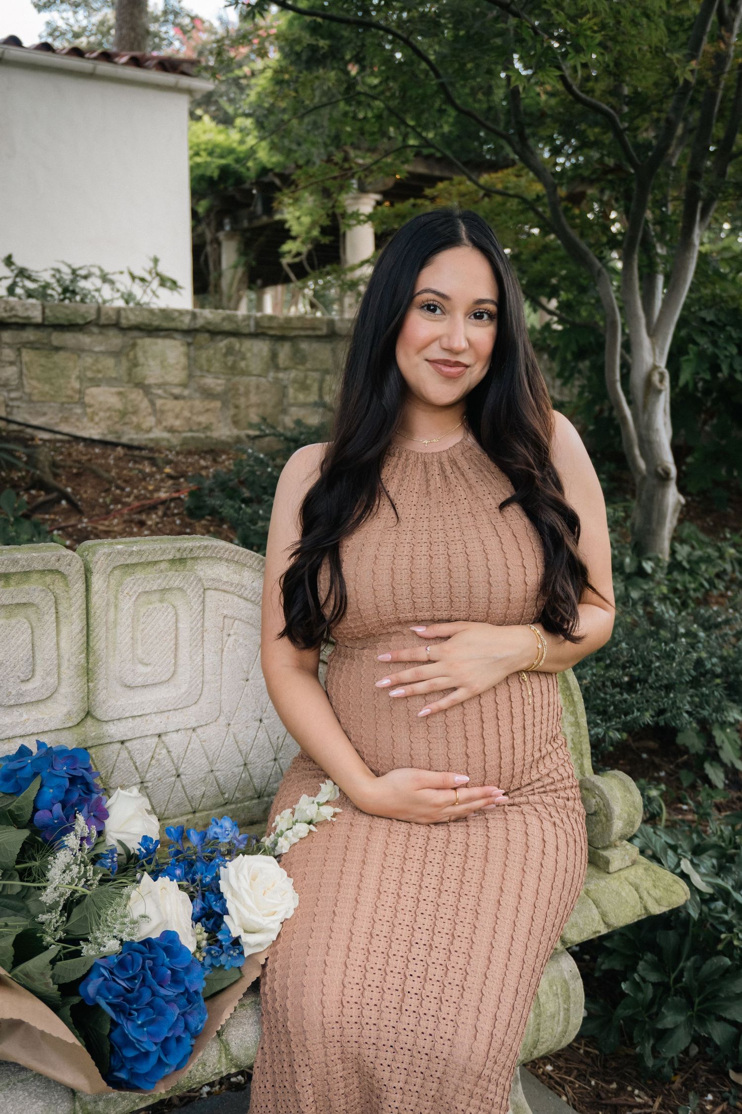 Expectant mother in beige knit dress holds her baby bump while posing with blue and white flowers in a garden setting.