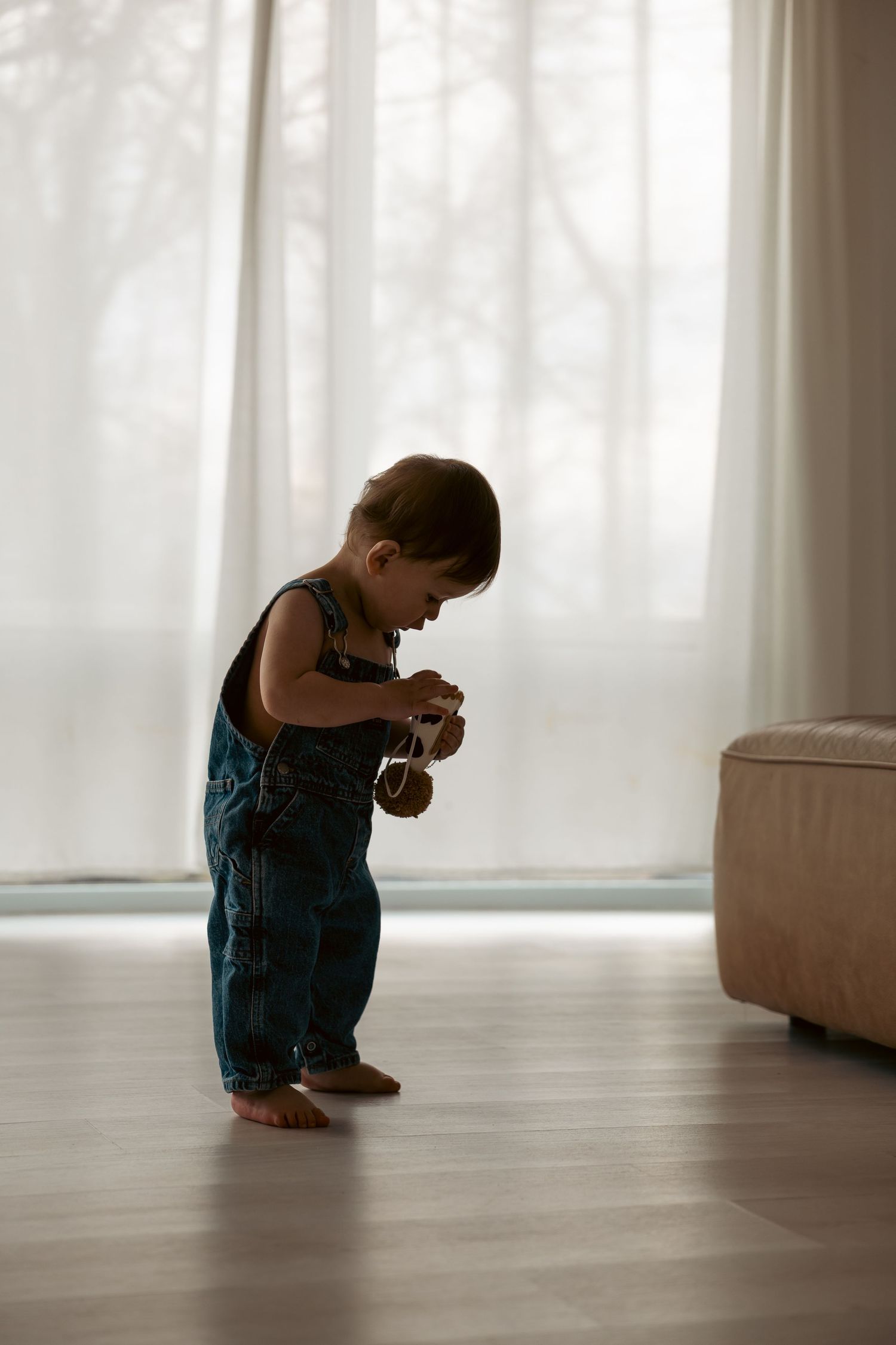 Small child in teal dress sits crouched down on hardwood floor near white walls in bright sunlit room.