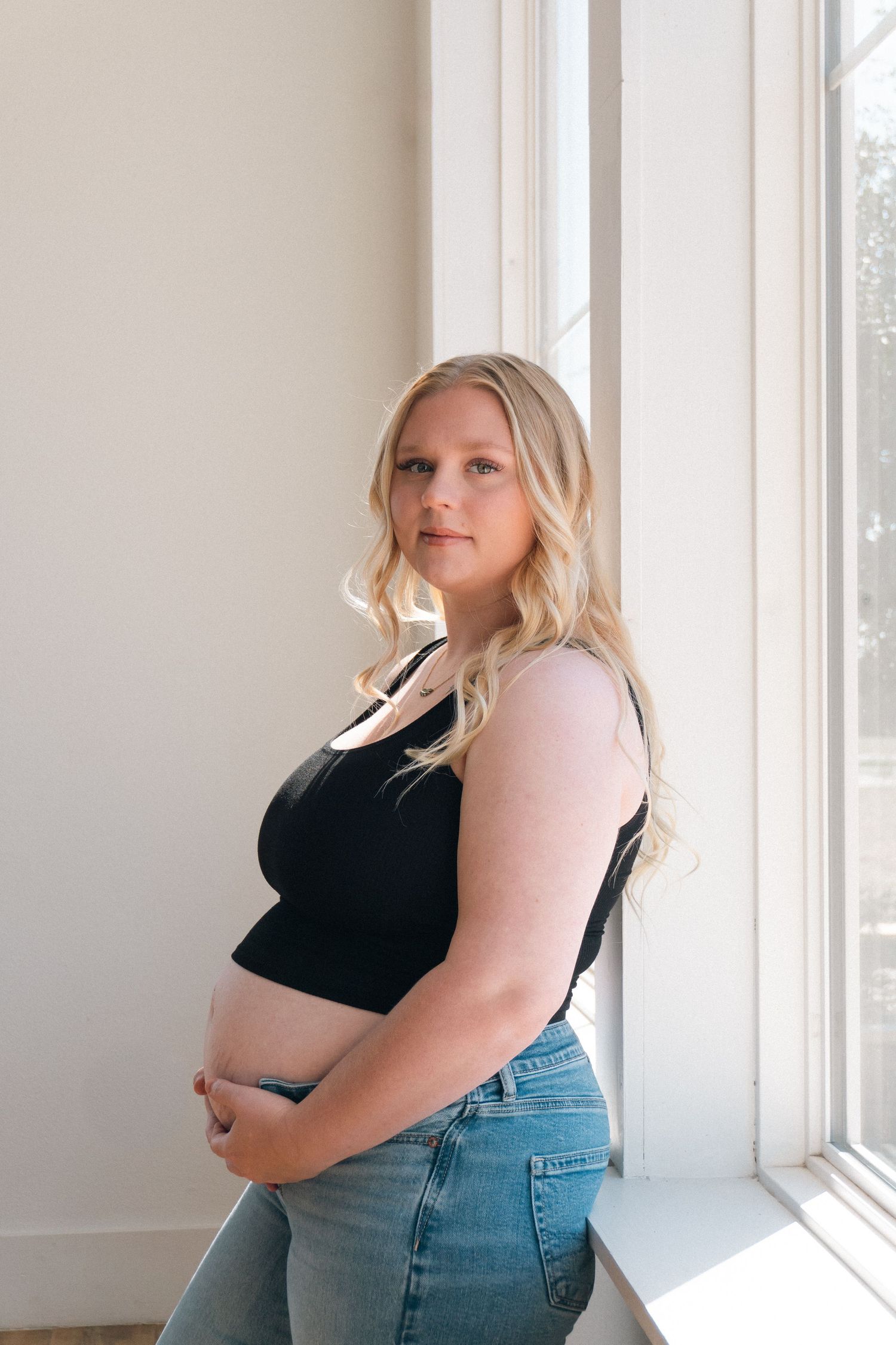 A maternity portrait by a window featuring a woman in casual attire.