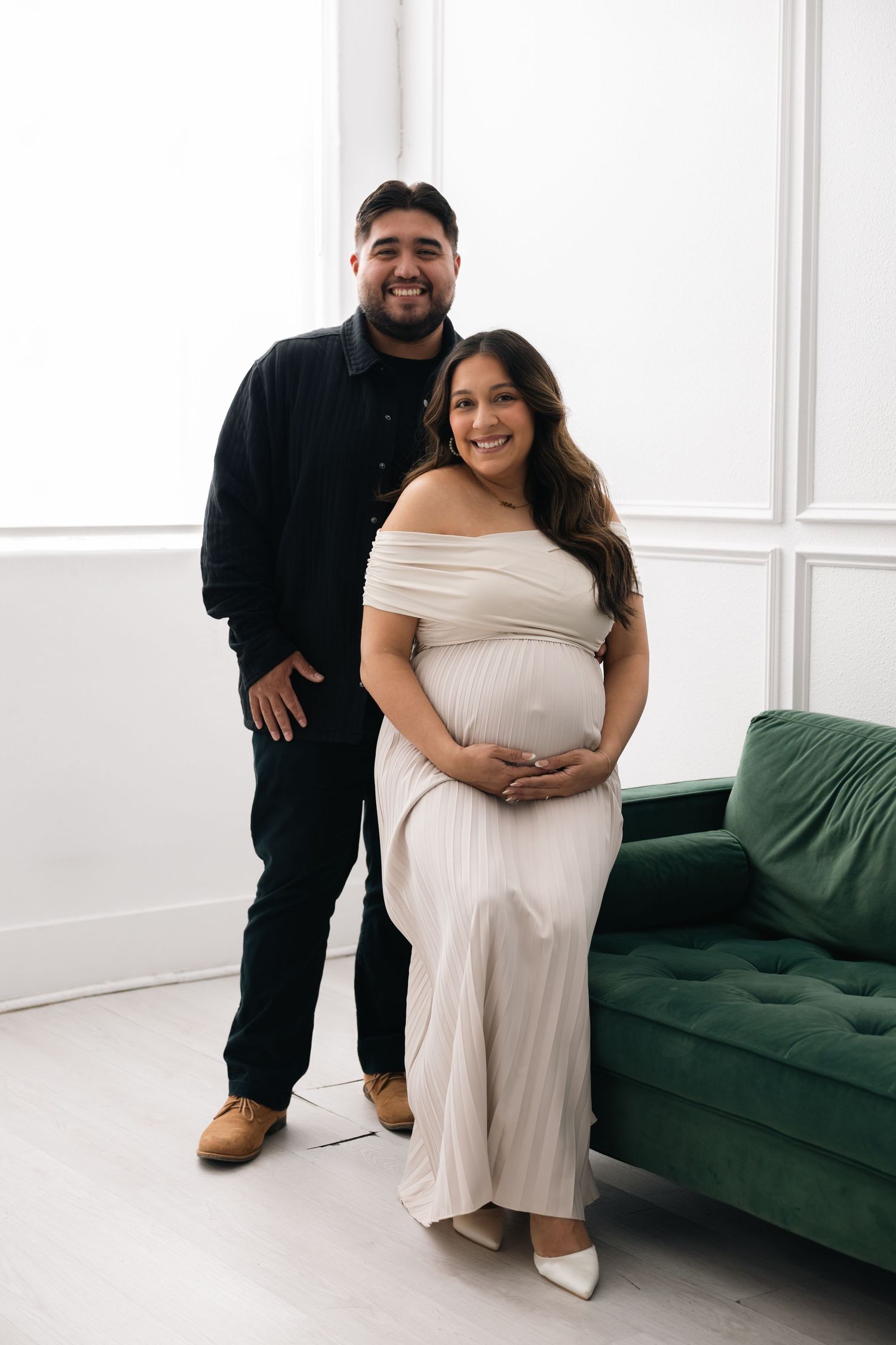 A couple poses together in a white studio with a green velvet couch, the woman wearing an elegant off-shoulder maternity dress.