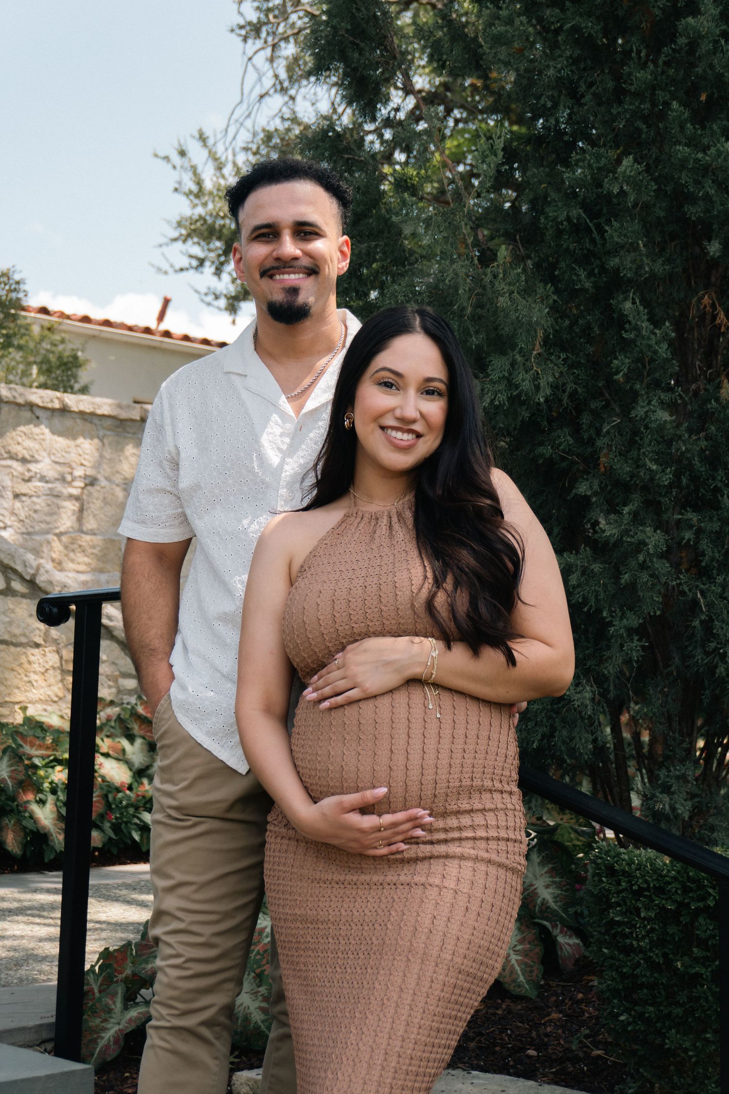 A couple poses together outdoors in coordinated outfits, with the woman wearing a tan knit dress against a natural backdrop.