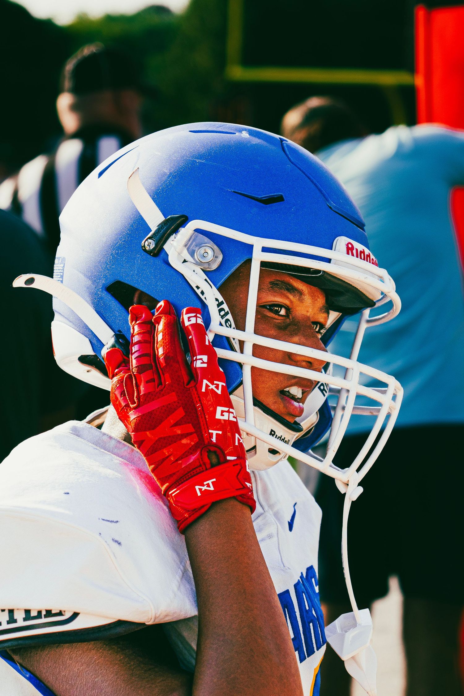 Young football player in blue Karns helmet and red gloves adjusting his helmet on the sideline.