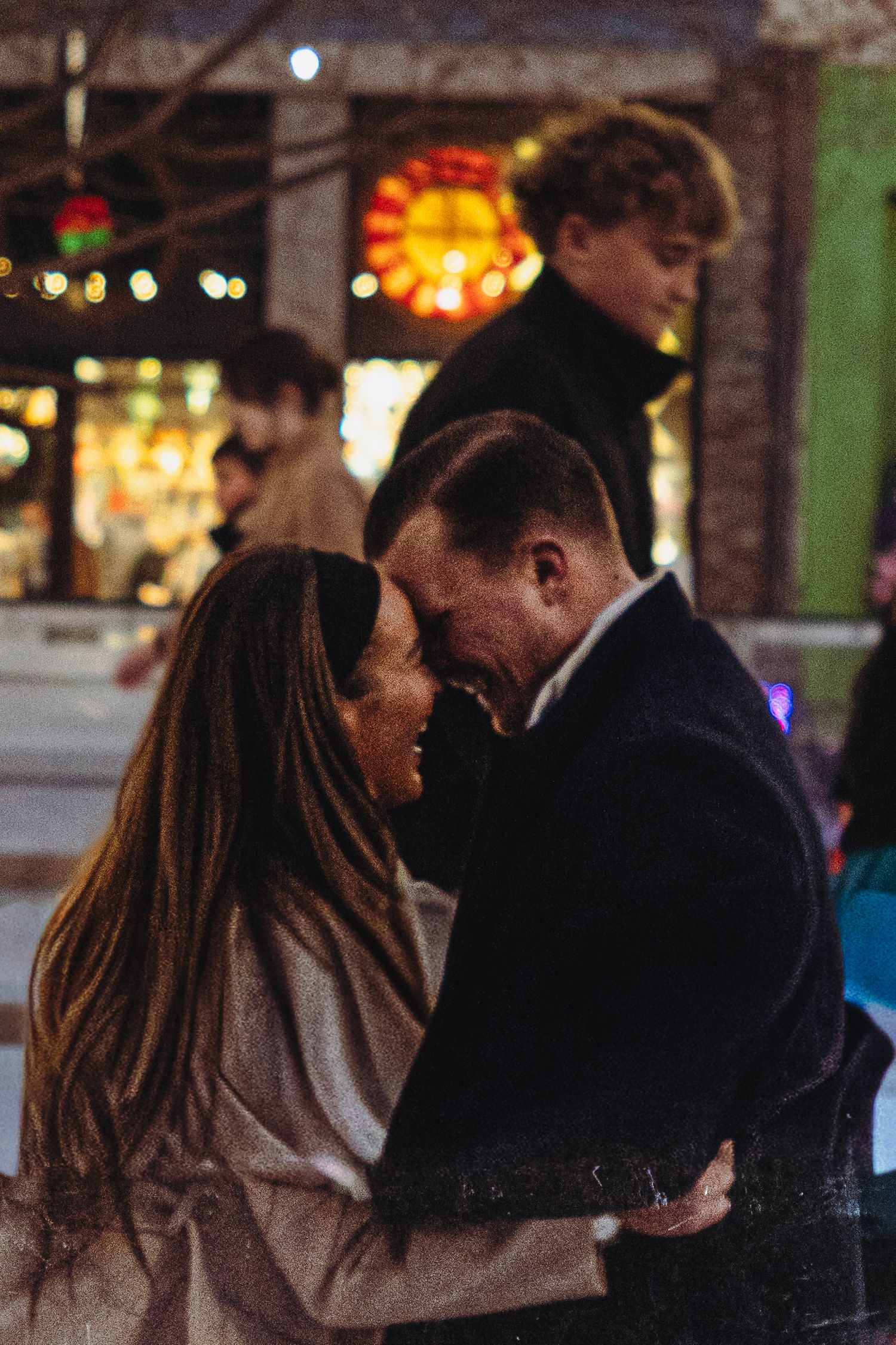 Romantic couple embracing and nearly kissing at a festive outdoor ice skating rink at night.