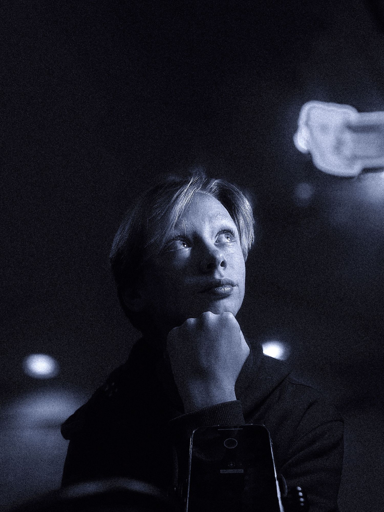 Moody black and white photo of a woman resting her chin on her hand, gazing upward in a dark setting.