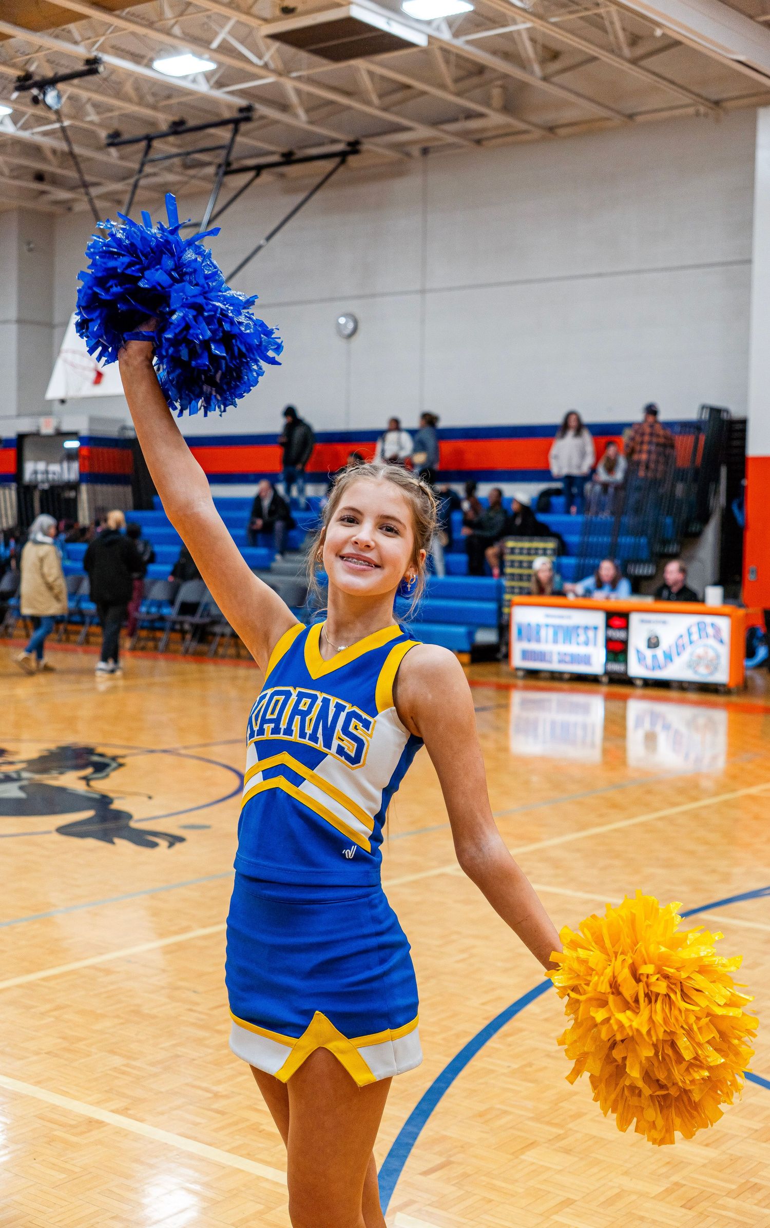Smiling cheerleader in blue and gold uniform holds pom-poms during an indoor basketball game.
