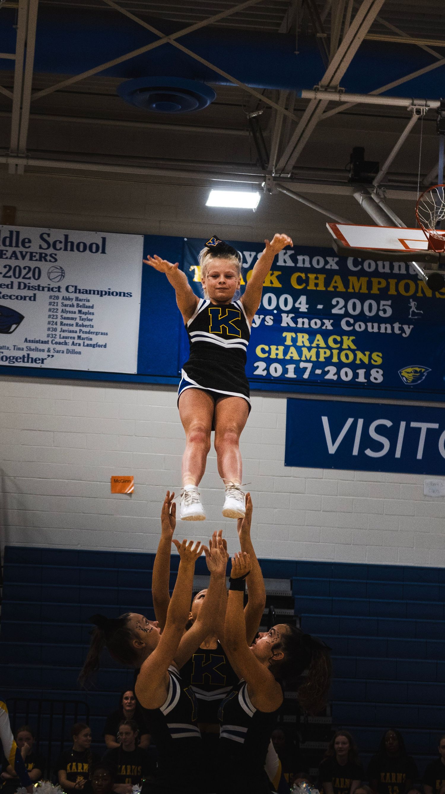 Cheerleader being lifted high in a stunt by teammates in a gymnasium with championship banners on the wall.