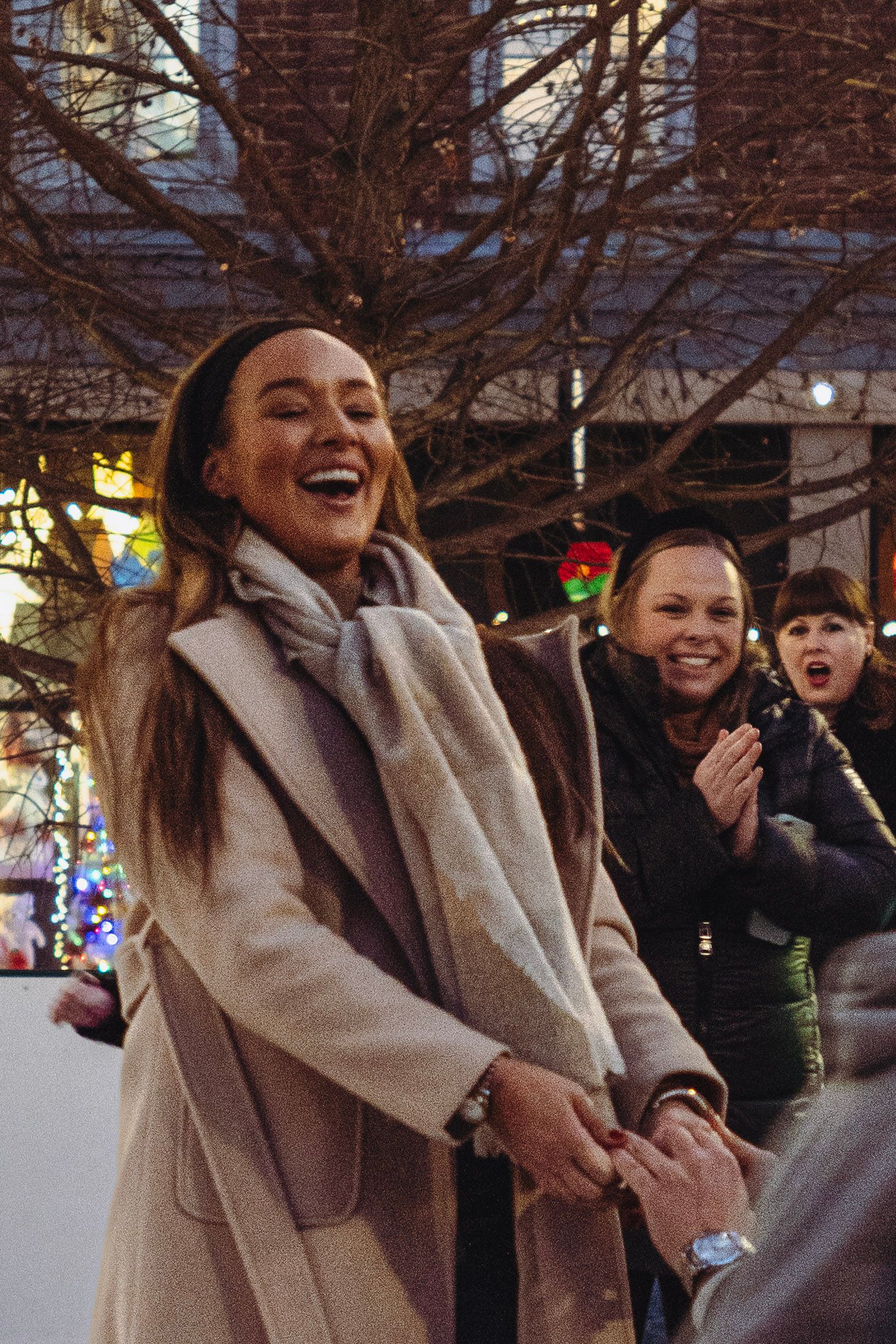 Joyful woman laughing outdoors at a festive ice skating rink while friends celebrate behind her.