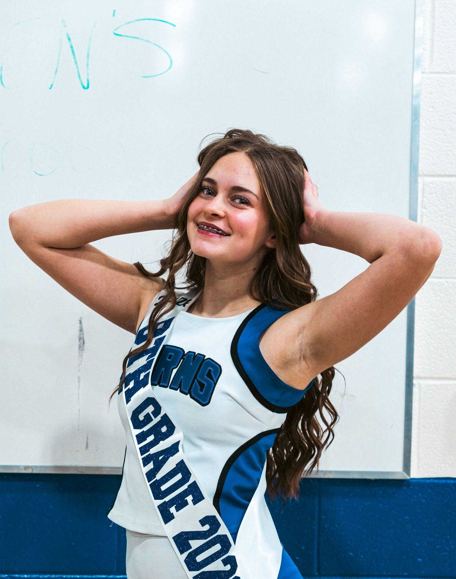 Young cheerleader in white and blue uniform posing with hands behind head, smiling confidently.