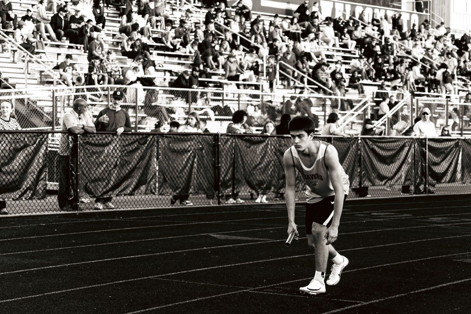 A track athlete crouches in starting position on a black track with packed stadium bleachers behind.