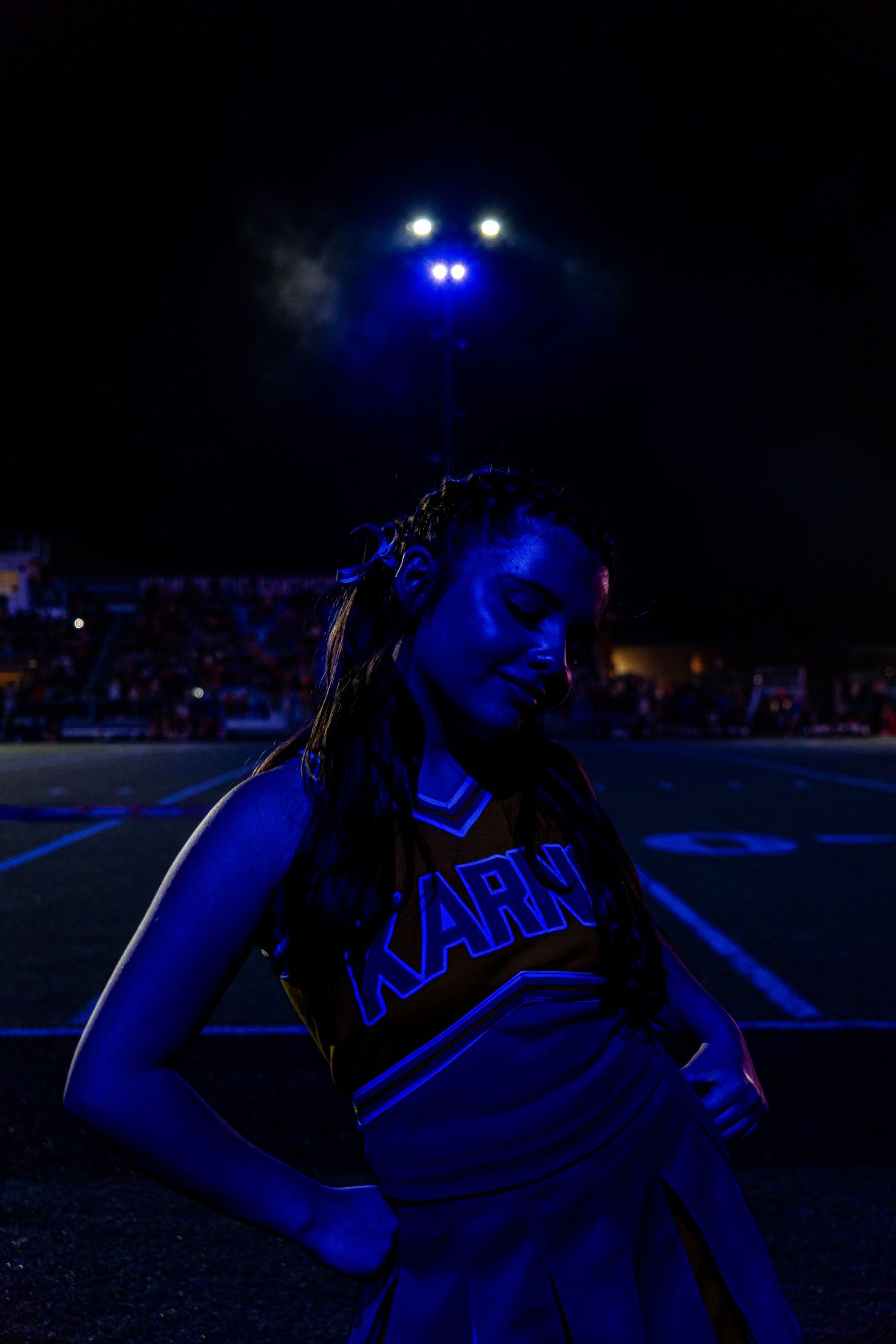 A Karns cheerleader poses dramatically under blue stadium lights during a night football game.