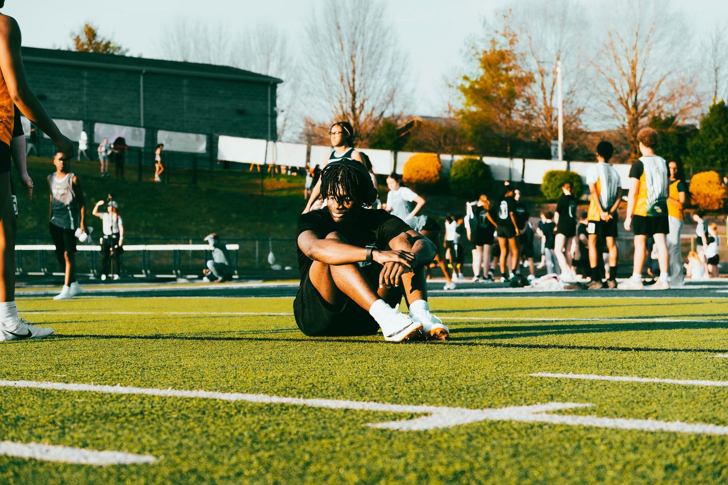 A track athlete sits on green turf tying shoes while teammates warm up in background.