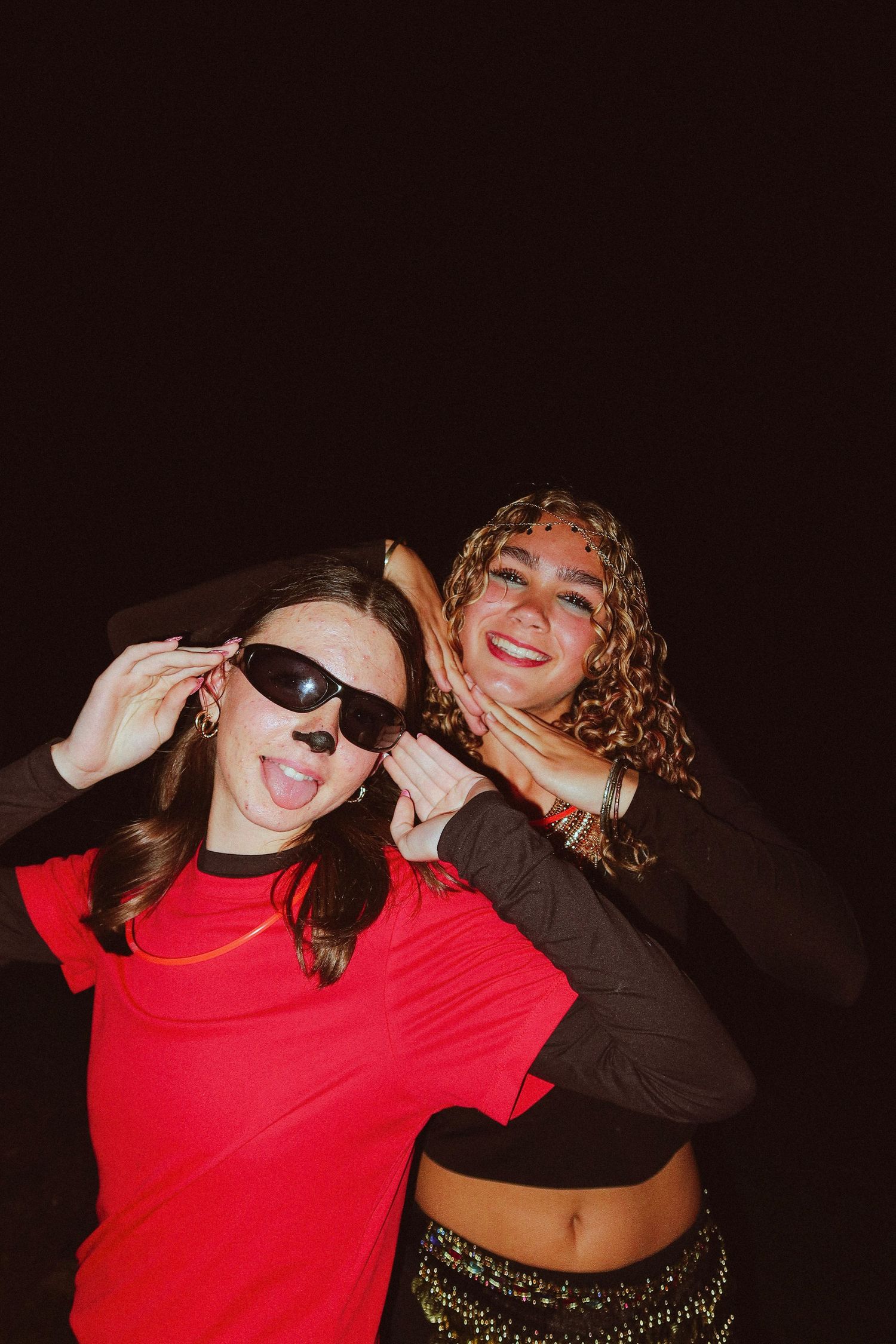 Two young women posing at night, one in red shirt with sunglasses, one in black crop top.