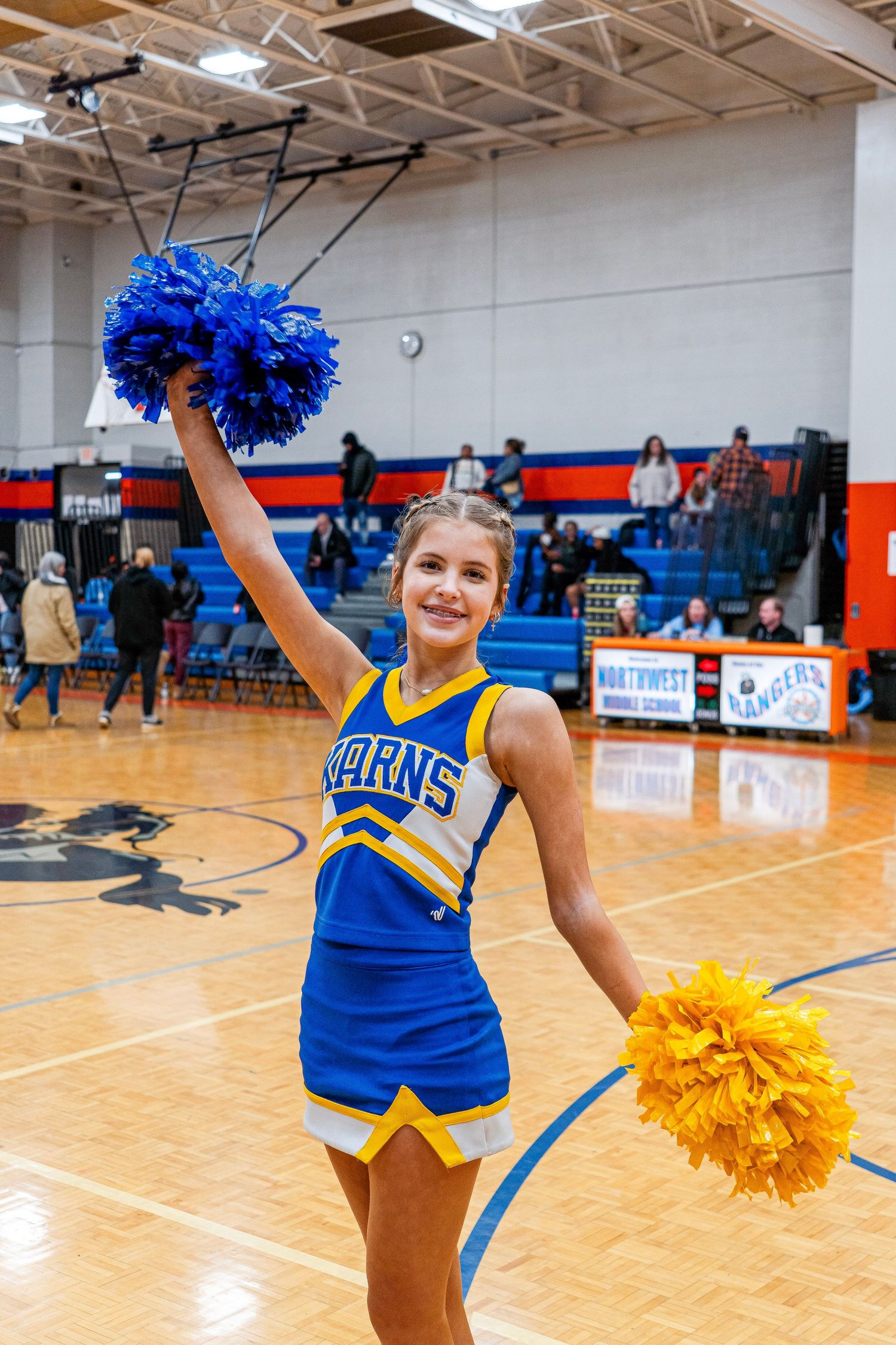 Karns cheerleader performing on gymnasium floor holding blue and gold pom-poms at a basketball game.