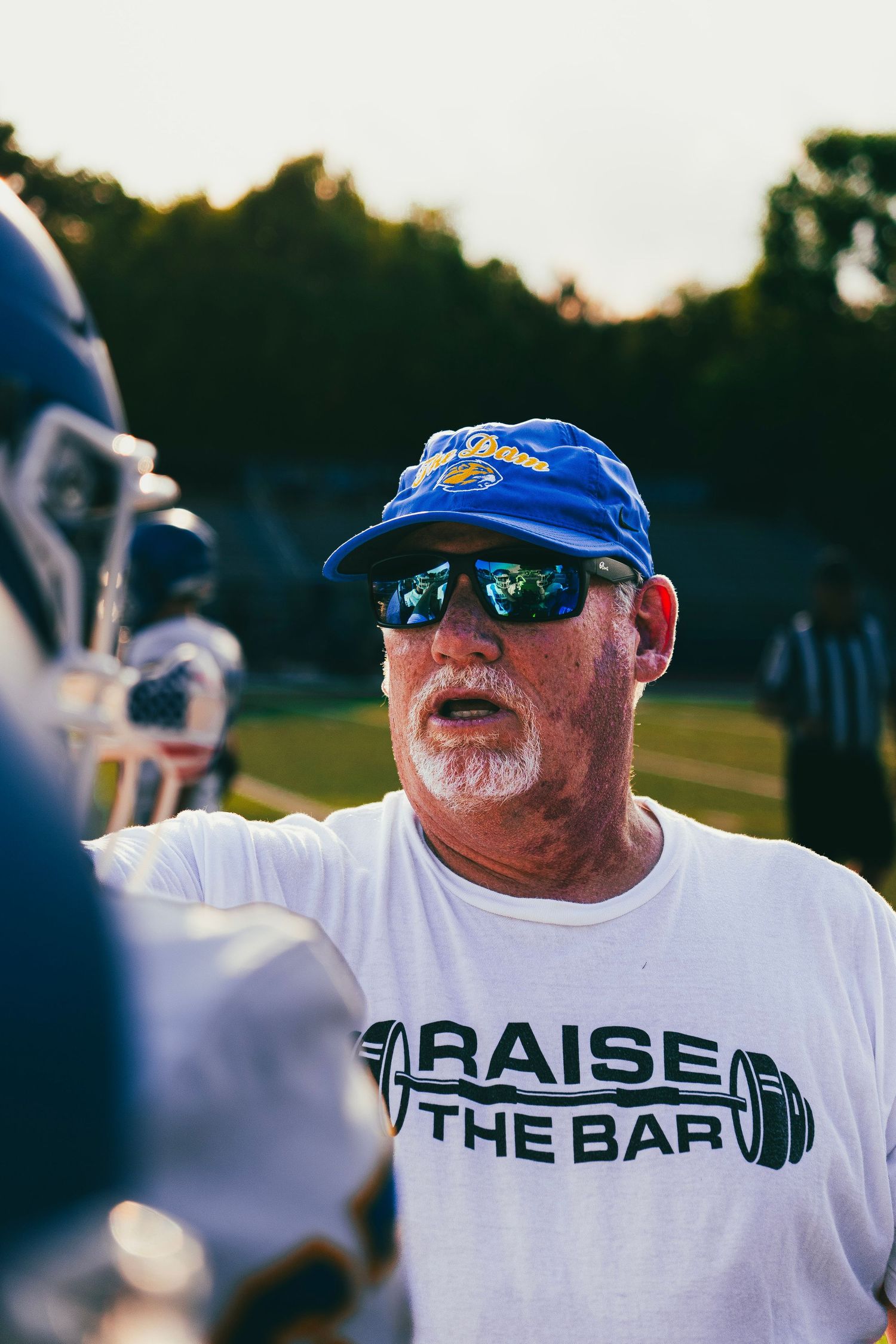 Football coach in blue cap and Raise The Bar shirt talks to players on sideline.