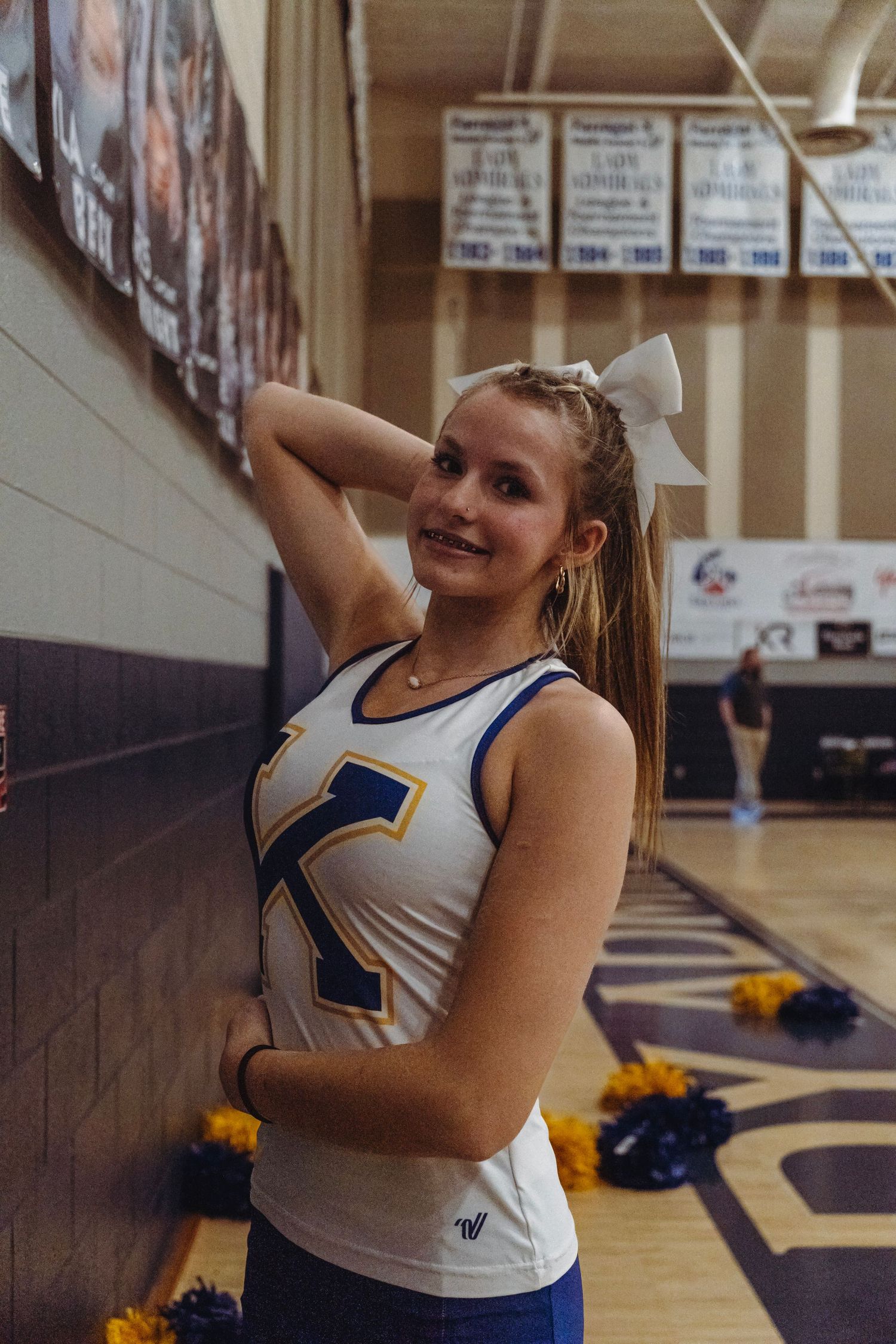 High school cheerleader in white and blue uniform poses in a gymnasium before a game.