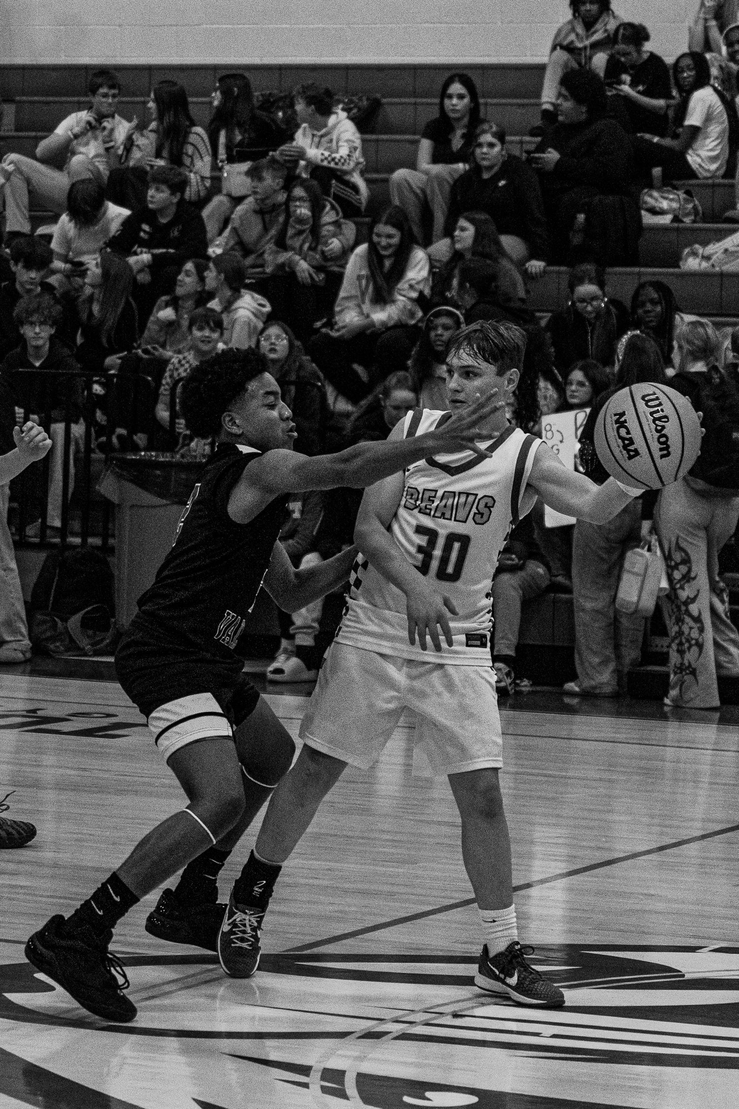 High school basketball player #30 shields ball from defender during packed gym game.