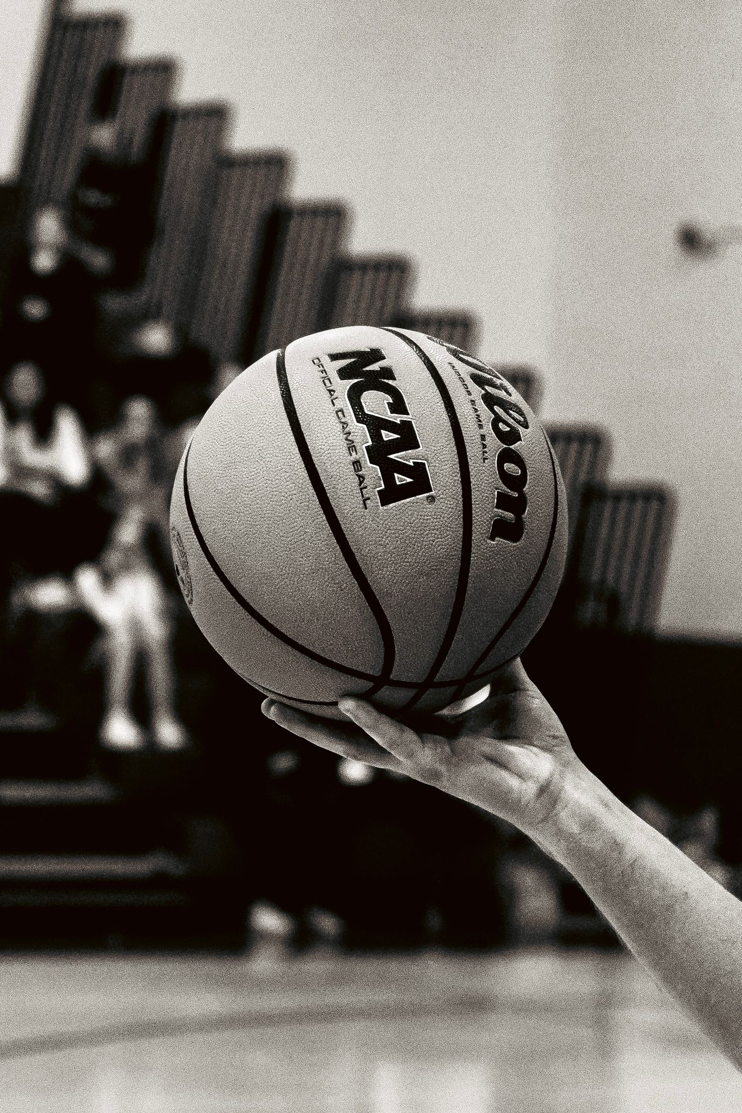 Hand holding a Wilson NCAA official basketball aloft in black and white with blurred bleachers in background.