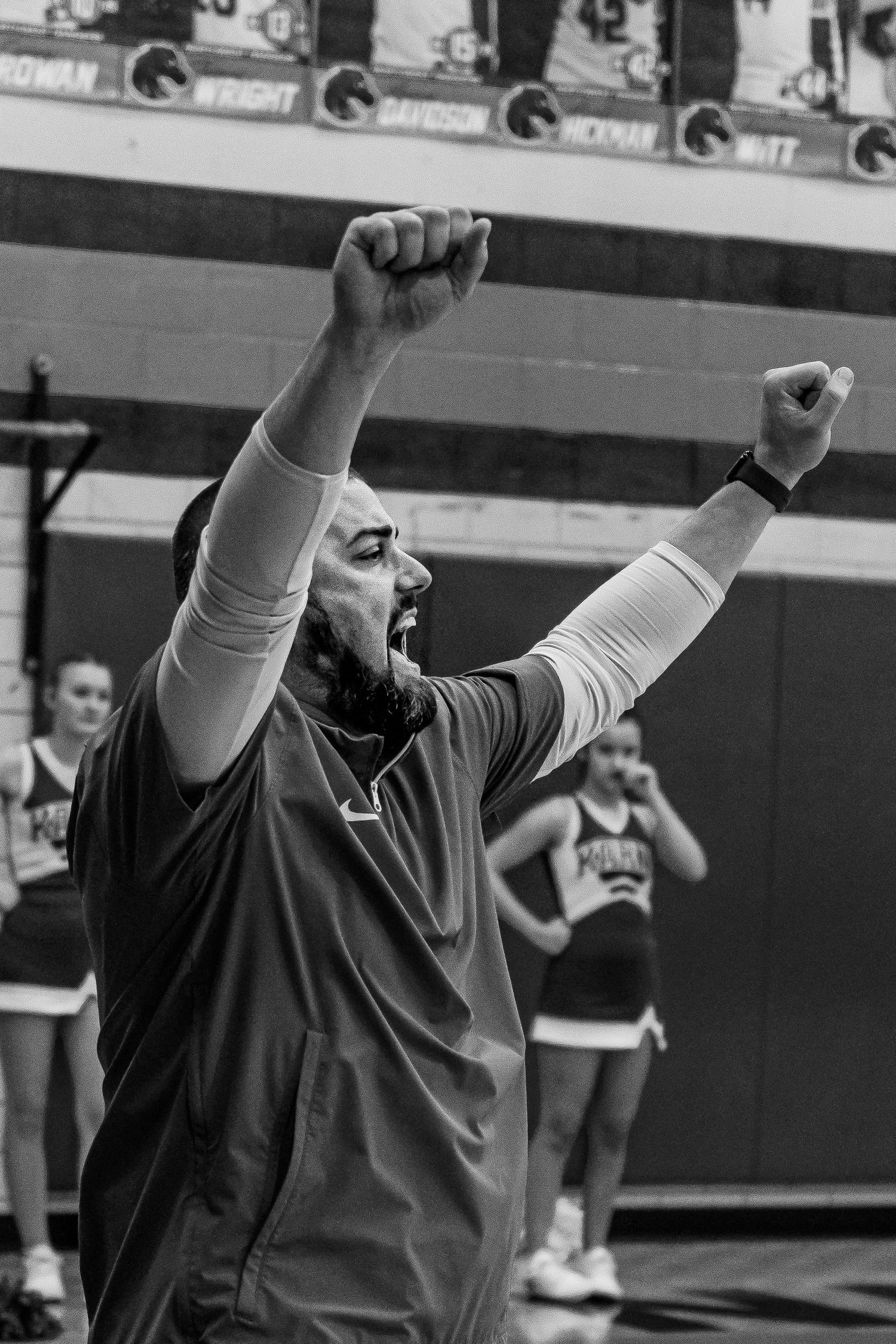 Bearded male basketball coach celebrates with fists raised in a black and white gym photo.