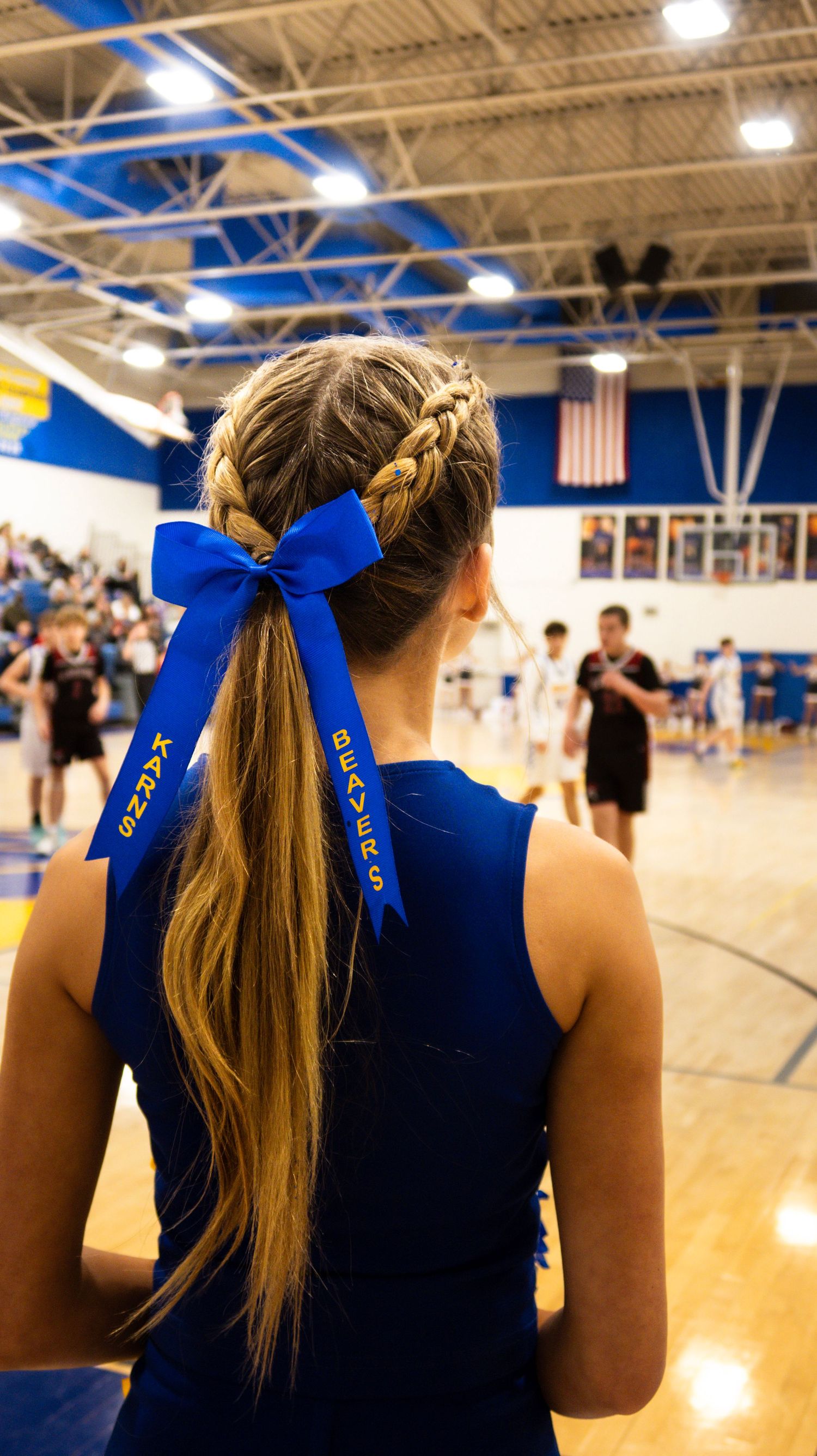 Back view of cheerleader with braided ponytail and blue Beavers bow in school gym.