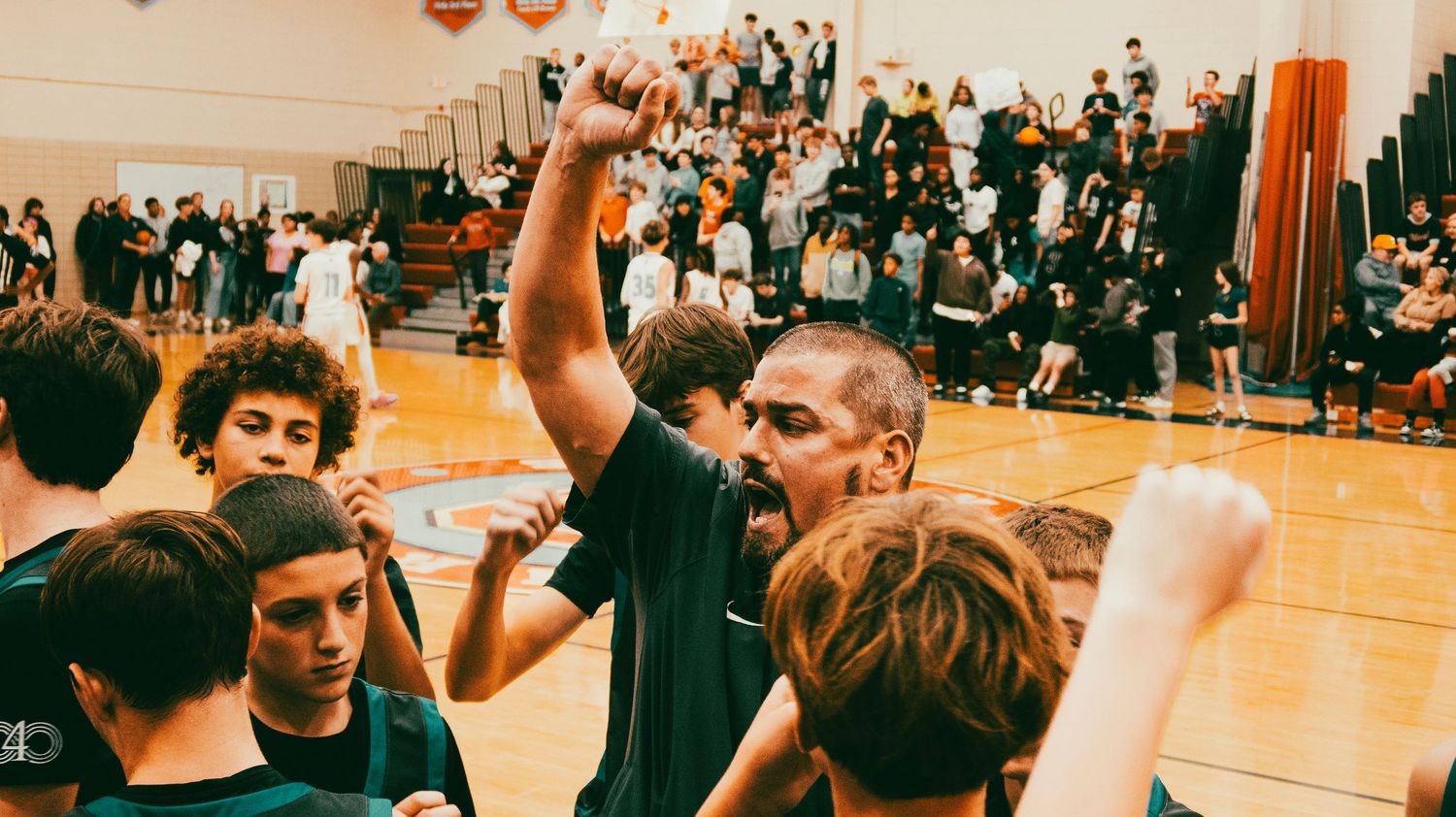 Energetic basketball coach raises fist celebrating with young players in crowded gymnasium after game.