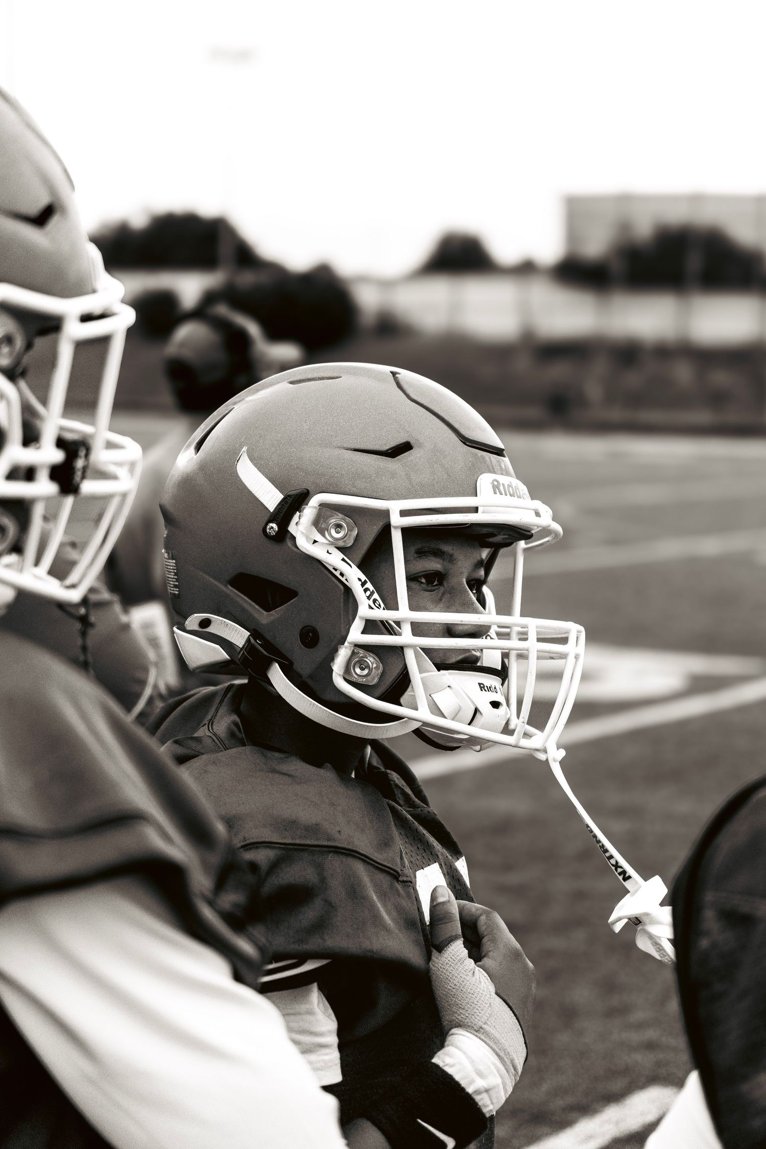 Black and white photo of young football player in helmet and pads on sideline.