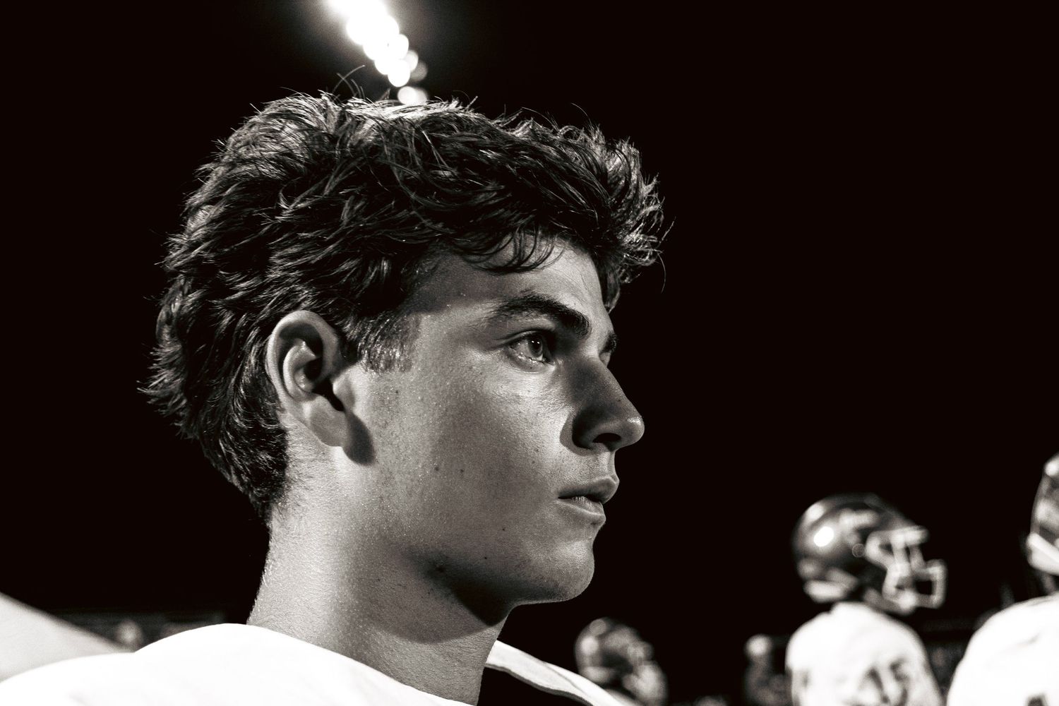 Black and white portrait of a young male high school football player on the sideline at night.