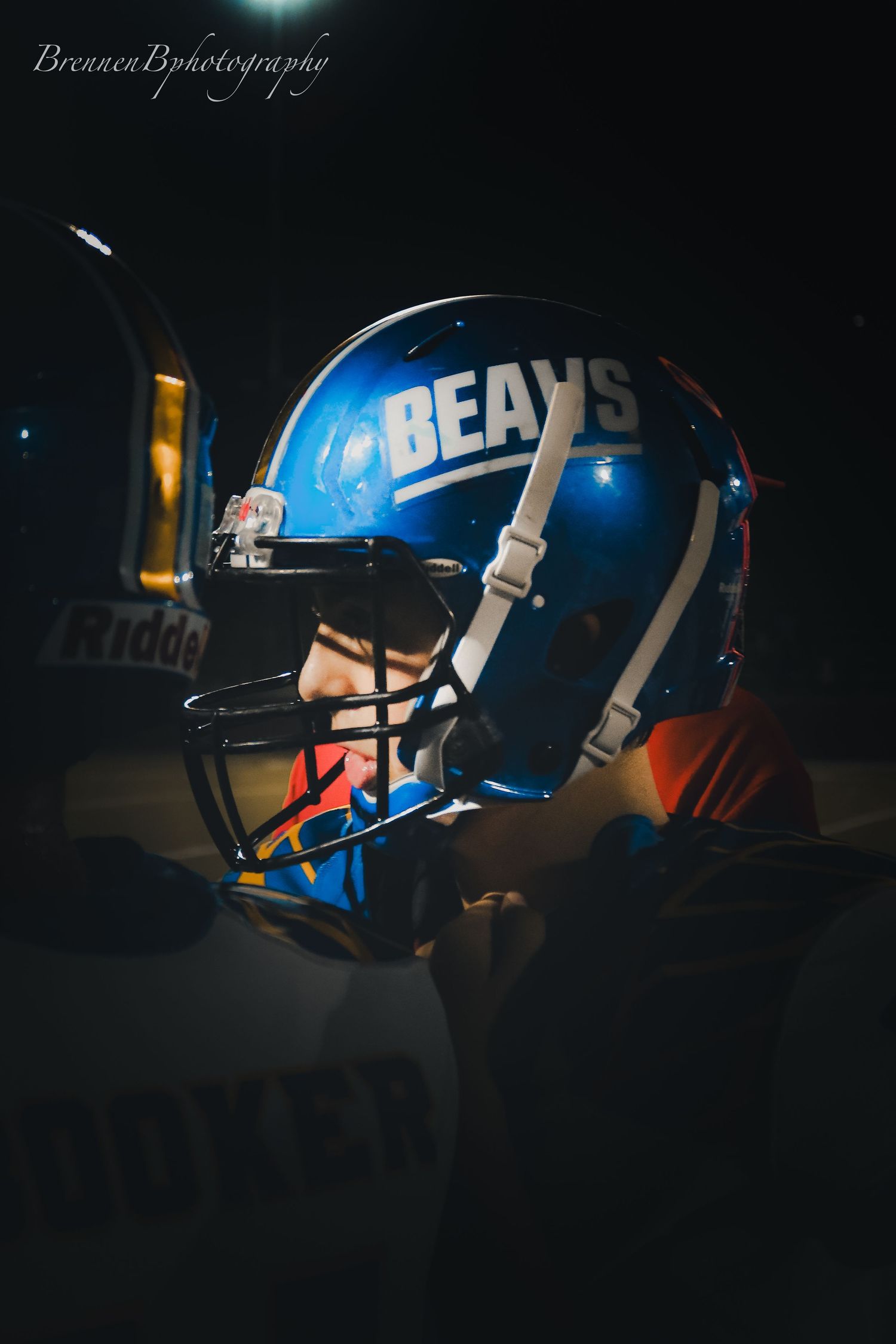 Dramatic close-up of a Beavs football player wearing a blue helmet illuminated against a dark background at night.