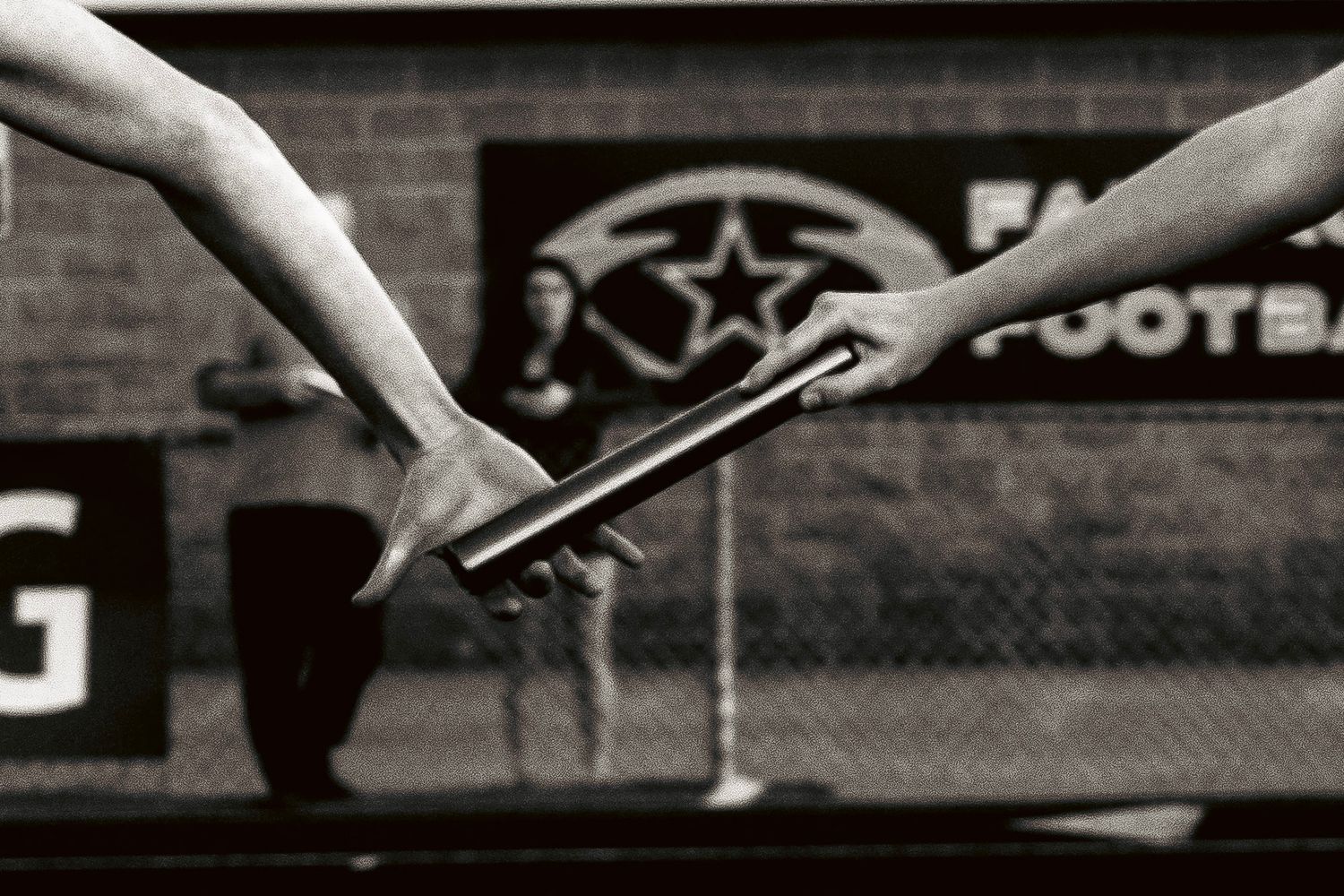 Black and white image of athletes passing a relay baton during a track race near a football field.